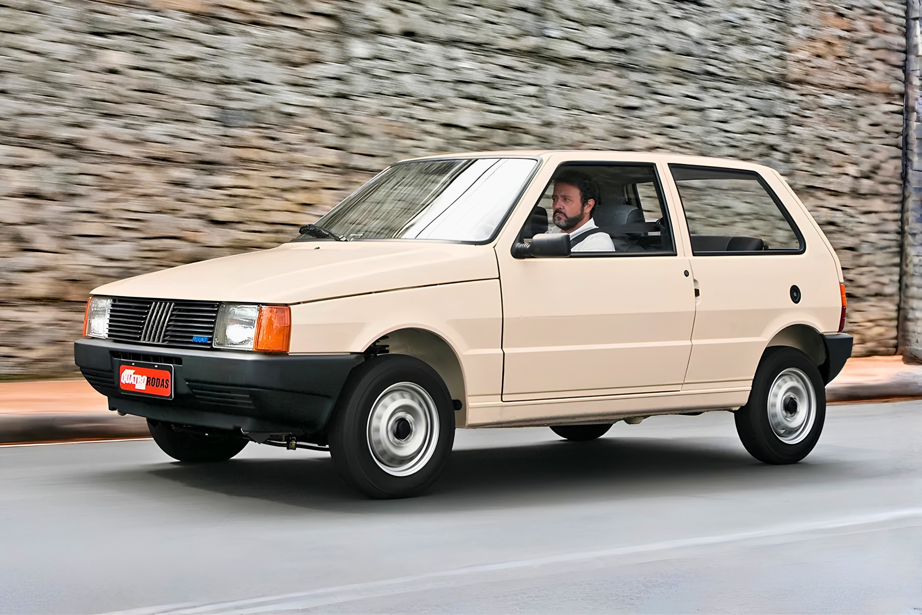 Um homem barbudo dirige um Fiat Uno bege, modelo antigo, em uma rua cinza com um muro de pedras ao fundo. O carro está em movimento, com o motorista visível pela janela.