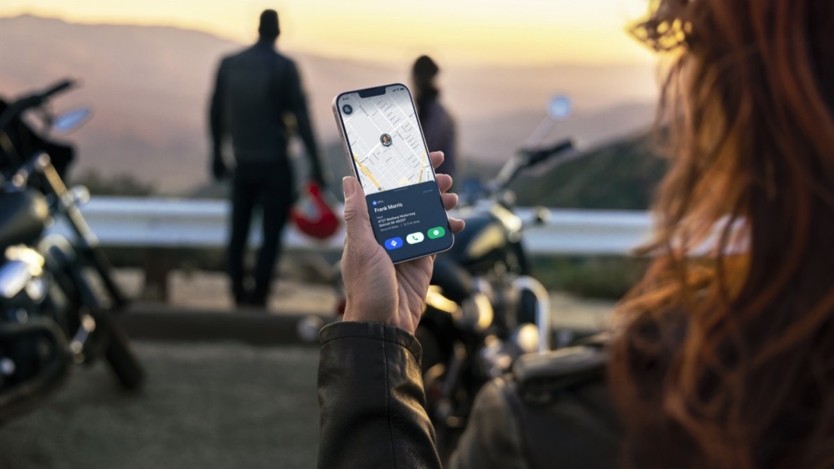 M&atilde;o de uma pessoa com jaqueta de couro segurando um smartphone com mapa e informa&ccedil;&otilde;es de contato, com motociclistas e motos desfocados ao fundo em um p&ocirc;r do sol