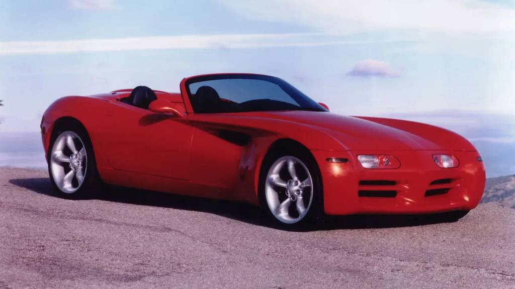 Carro esportivo convers&iacute;vel vermelho brilhante, com capota abaixada, estacionado em uma estrada asfaltada. O ve&iacute;culo tem rodas prateadas e design aerodin&acirc;mico. Ao fundo, um c&eacute;u azul claro com nuvens esparsas e uma paisagem montanhosa desfocada