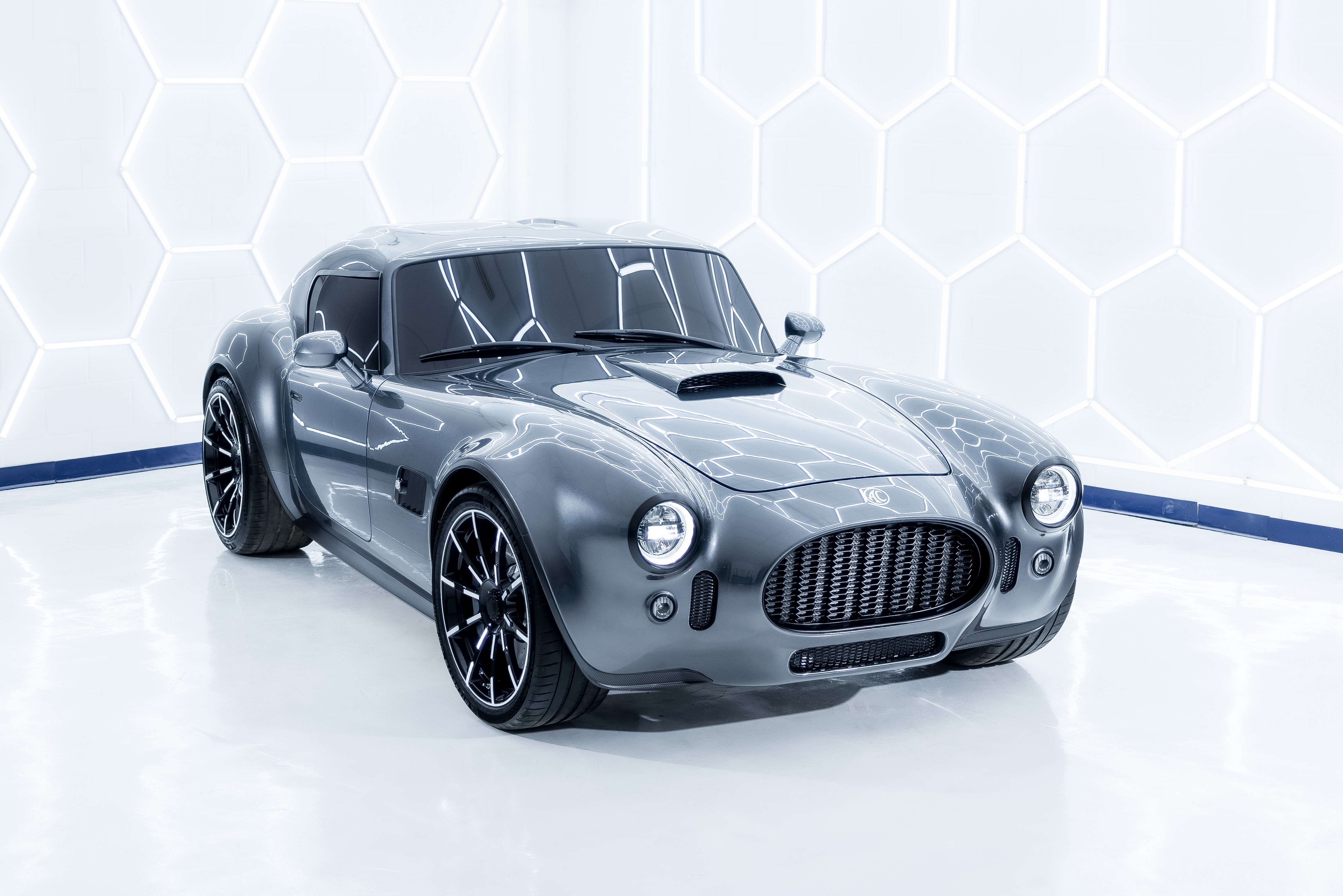 Carro esportivo cinza escuro, estilo cl&aacute;ssico, com cap&ocirc; longo e entrada de ar, rodas pretas e detalhes cromados, em um est&uacute;dio branco com ilumina&ccedil;&atilde;o hexagonal na parede