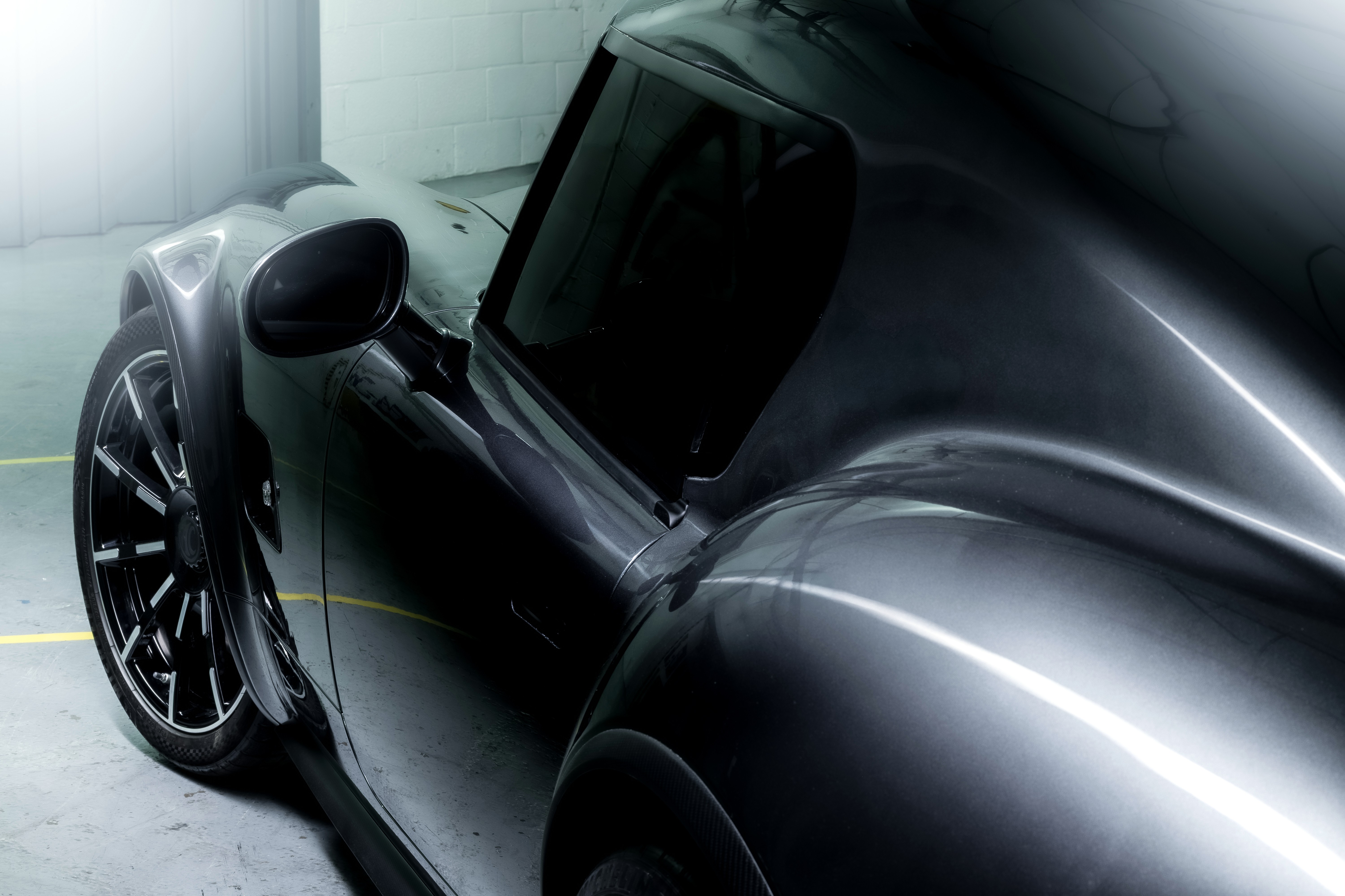 Lateral de um carro esportivo cinza escuro met&aacute;lico, com reflexos de luz branca e linhas curvas. A roda dianteira preta com detalhes prateados est&aacute; vis&iacute;vel, e o retrovisor lateral aponta para frente. O carro est&aacute; em um ambiente interno com paredes de tijolos brancos e piso cinza com uma linha amarela