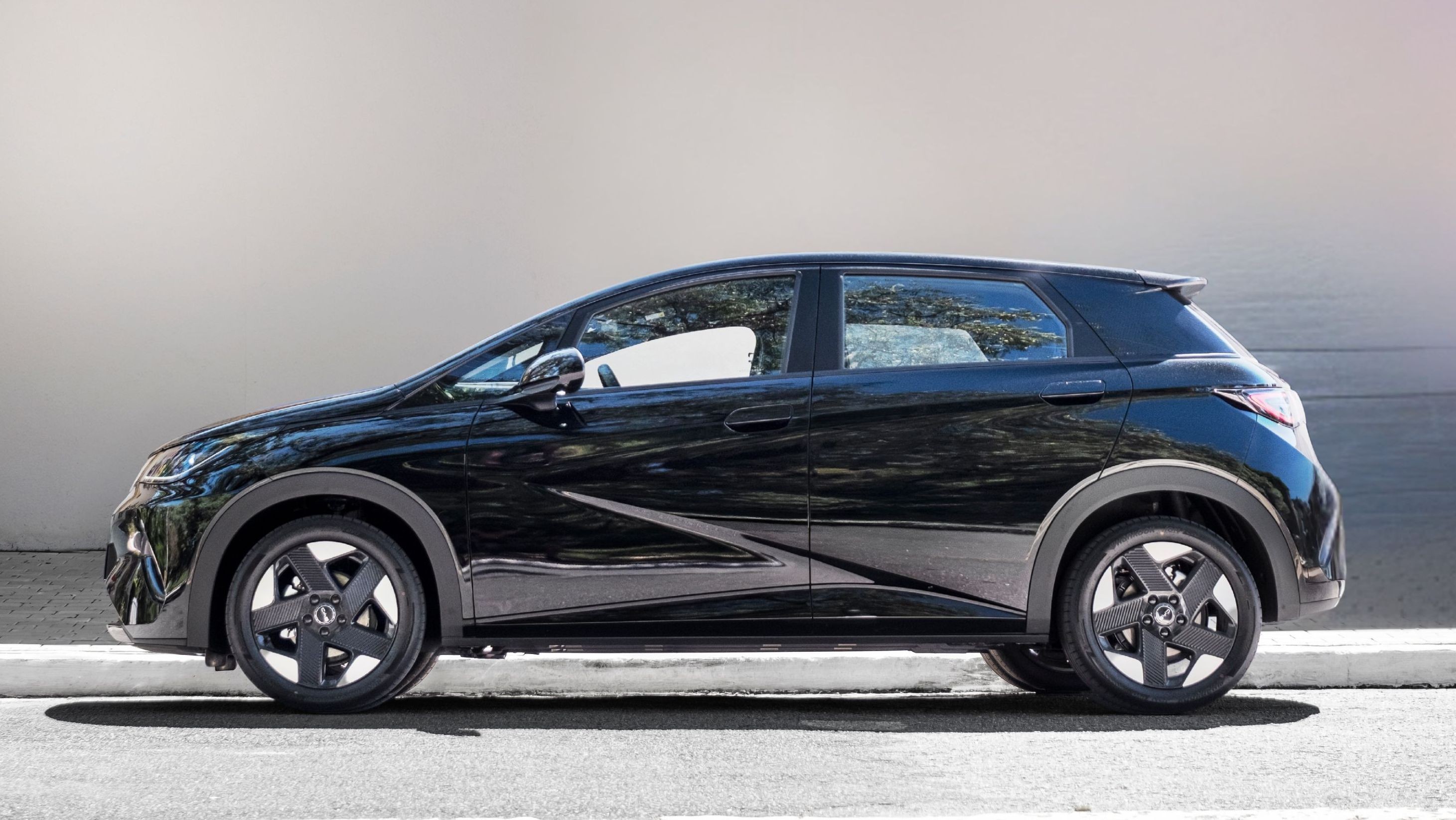 Carro preto compacto com detalhes cromados, rodas escuras de cinco raios, estacionado em asfalto cinza claro com meio-fio branco, sob luz solar forte e fundo cinza claro
