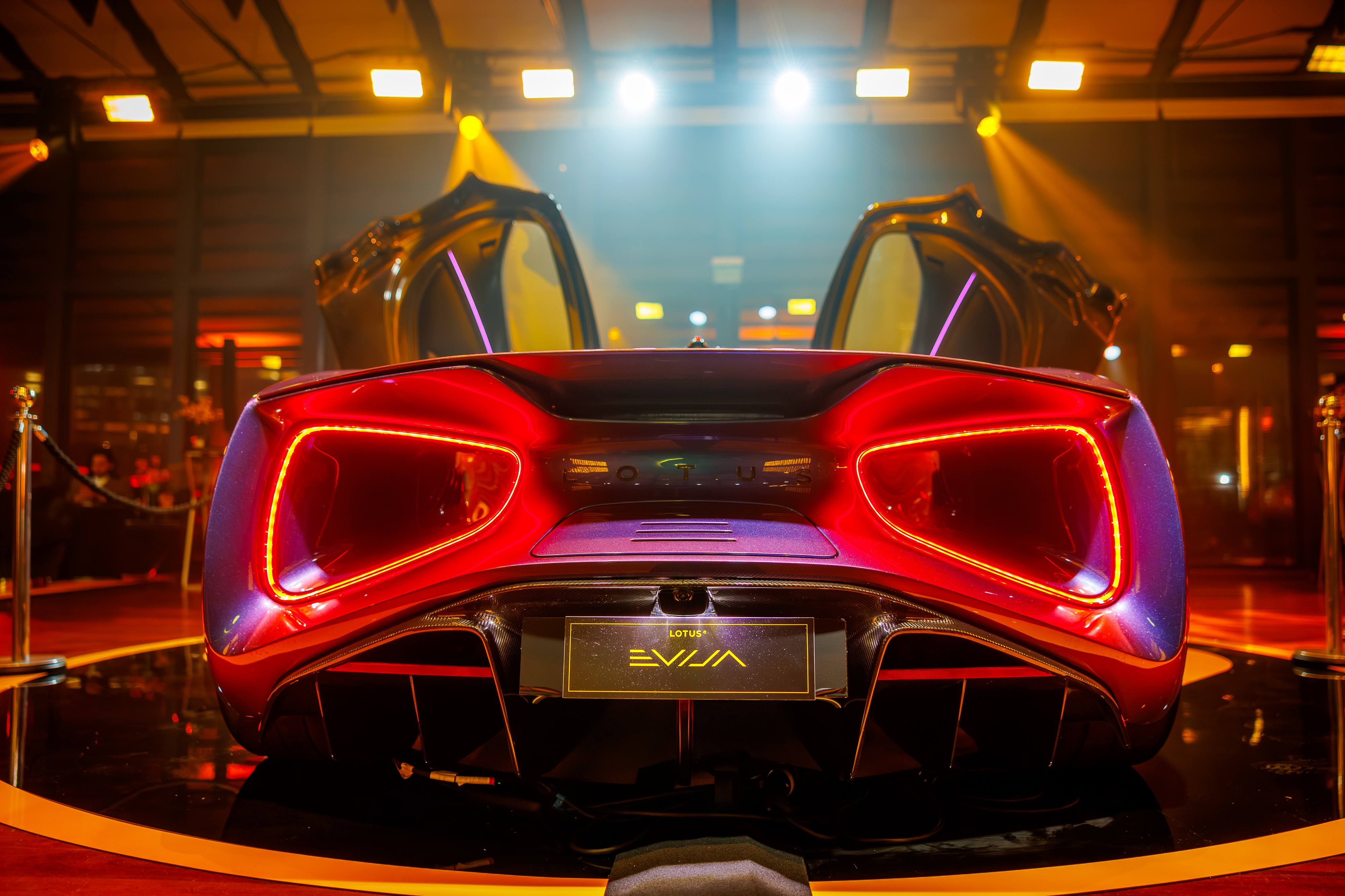 Traseira de um carro esportivo vermelho brilhante, modelo Lotus Evija, com as portas abertas para cima, exibindo luzes traseiras em neon e um difusor aerodin&acirc;mico. O ve&iacute;culo est&aacute; em um palco escuro com ilumina&ccedil;&atilde;o laranja e roxa, cercado por pilares dourados e cord&otilde;es de veludo, sugerindo um evento de lan&ccedil;amento ou exposi&ccedil;&atilde;o