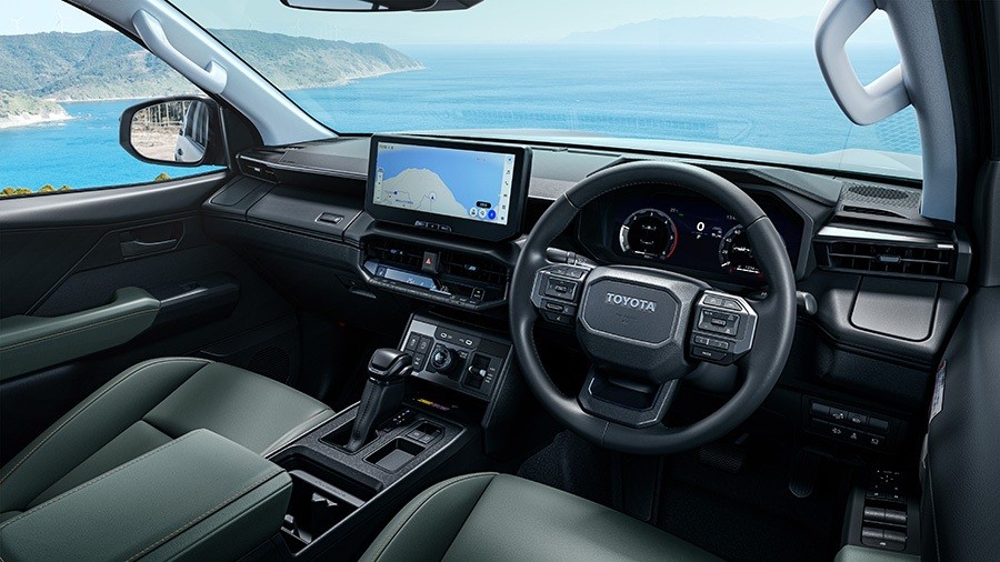 Interior de um carro Toyota moderno, com volante multifuncional, painel digital e tela multim&iacute;dia exibindo um mapa. Bancos e acabamentos em tons escuros, com detalhes em cinza. Ao fundo, uma paisagem costeira com mar azul e montanhas verdes, vista atrav&eacute;s do para-brisa