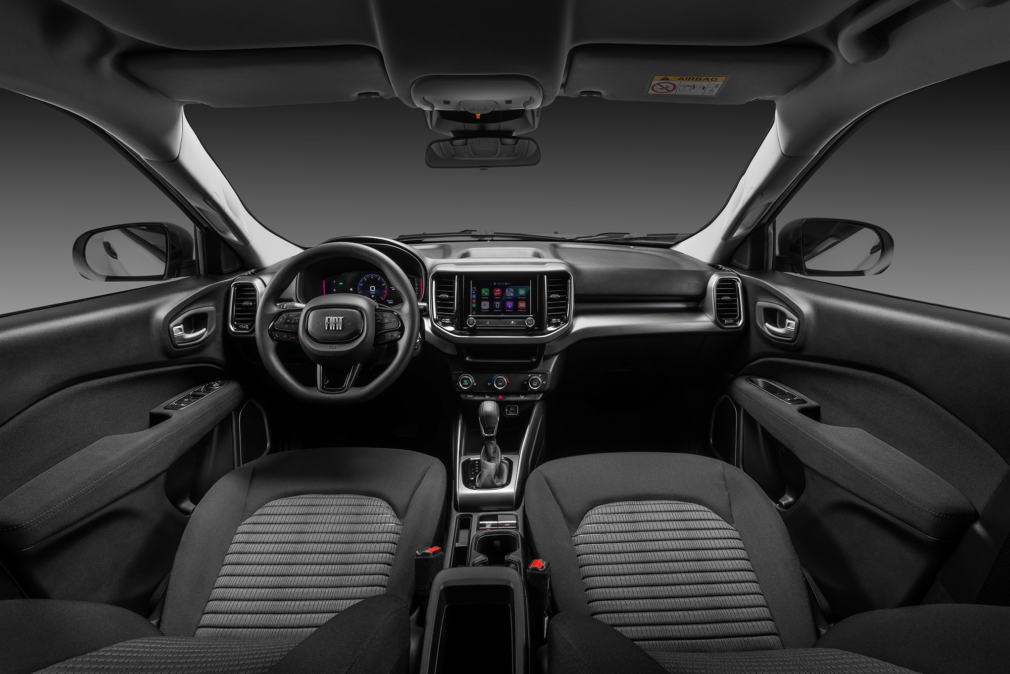 Interior de um carro moderno, com bancos dianteiros de tecido cinza e preto, painel escuro, volante com logo Fiat, tela multim&iacute;dia central e c&acirc;mbio autom&aacute;tico