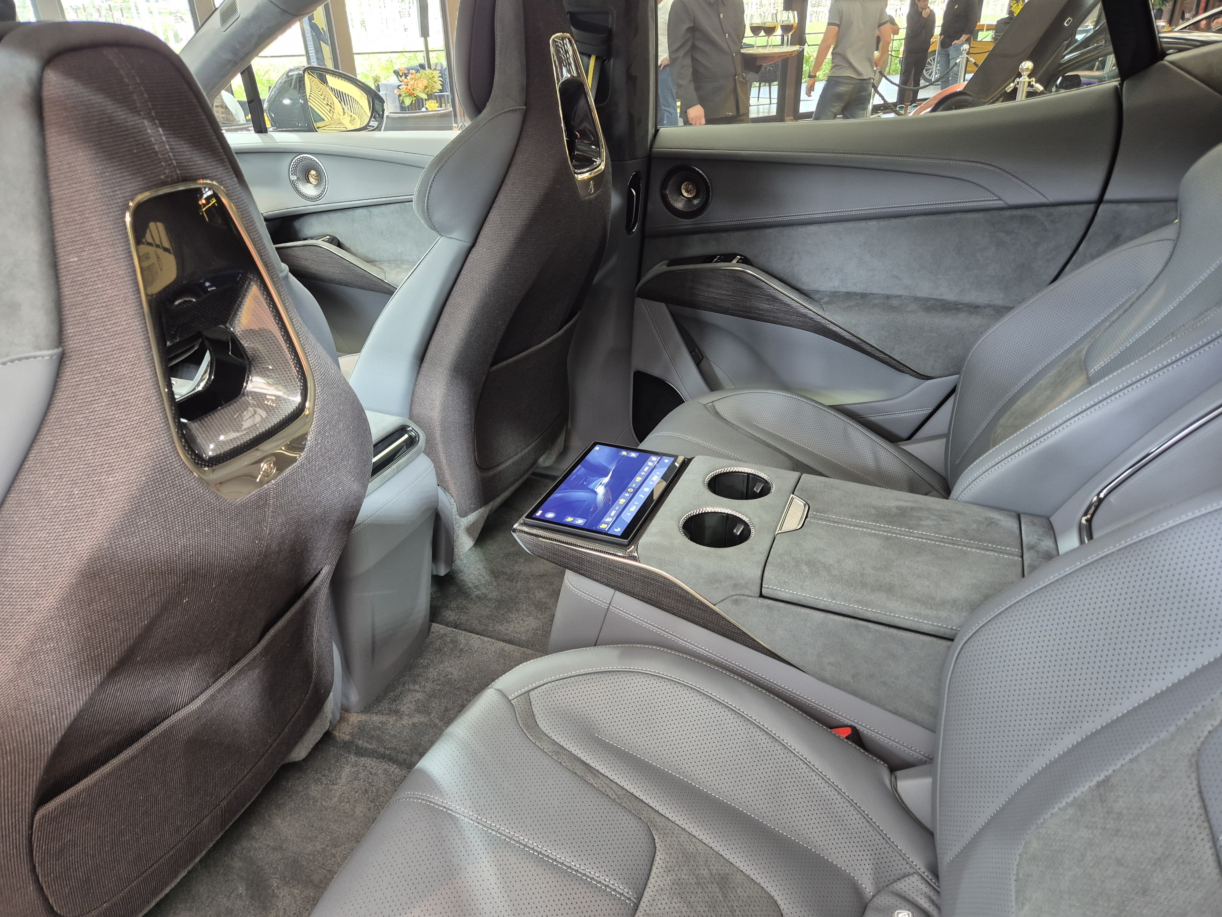 Interior de carro luxuoso com bancos de couro cinza e detalhes em camur&ccedil;a. Um tablet com tela azul est&aacute; no console central traseiro, ao lado de dois porta-copos. Pessoas desfocadas aparecem ao fundo, fora do ve&iacute;culo