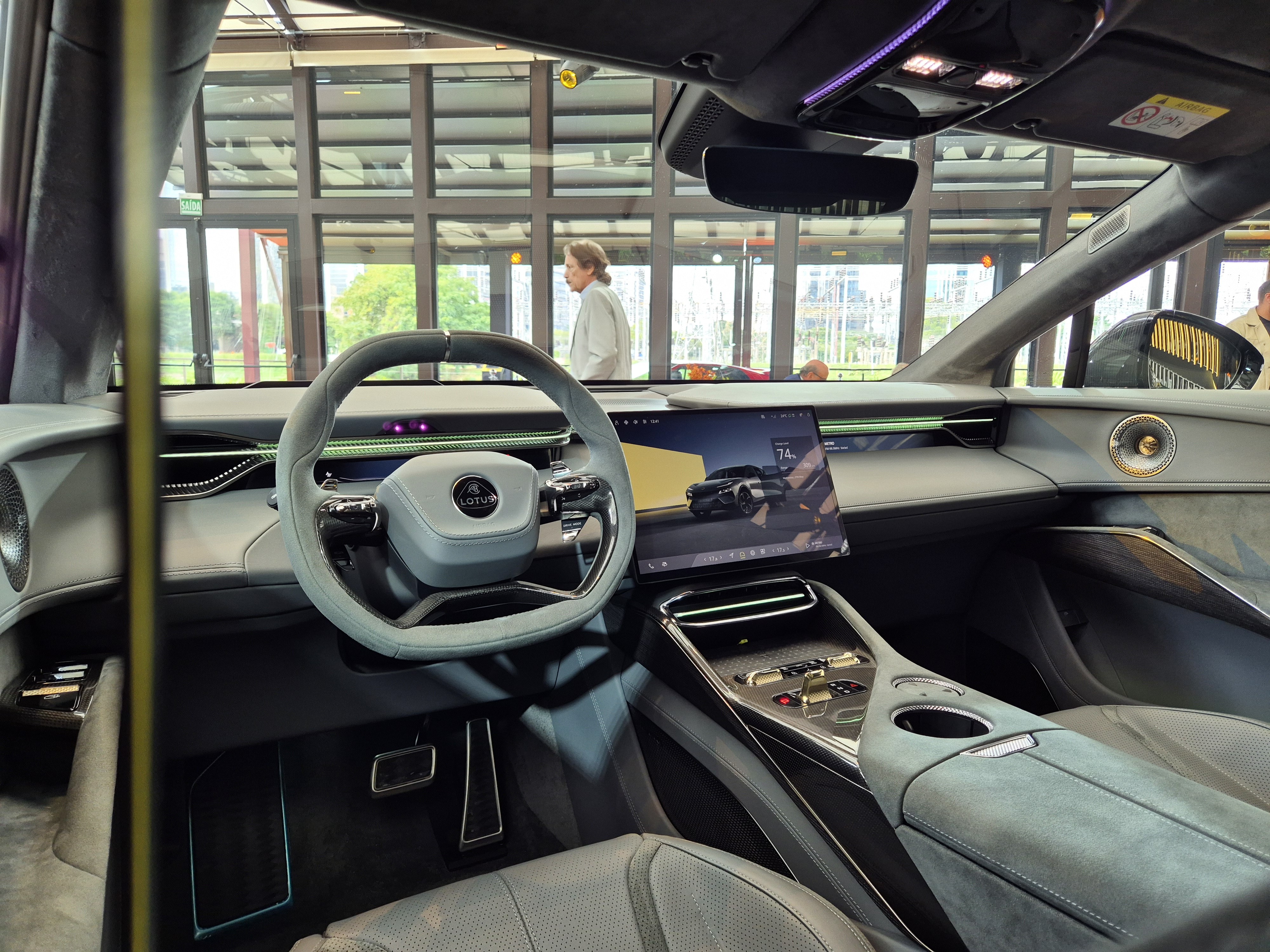 Interior de um carro Lotus cinza com volante esportivo, tela multim&iacute;dia exibindo outro carro e console central com bot&otilde;es e porta-copos. Uma pessoa de camisa clara caminha ao fundo, vis&iacute;vel atrav&eacute;s do para-brisa.