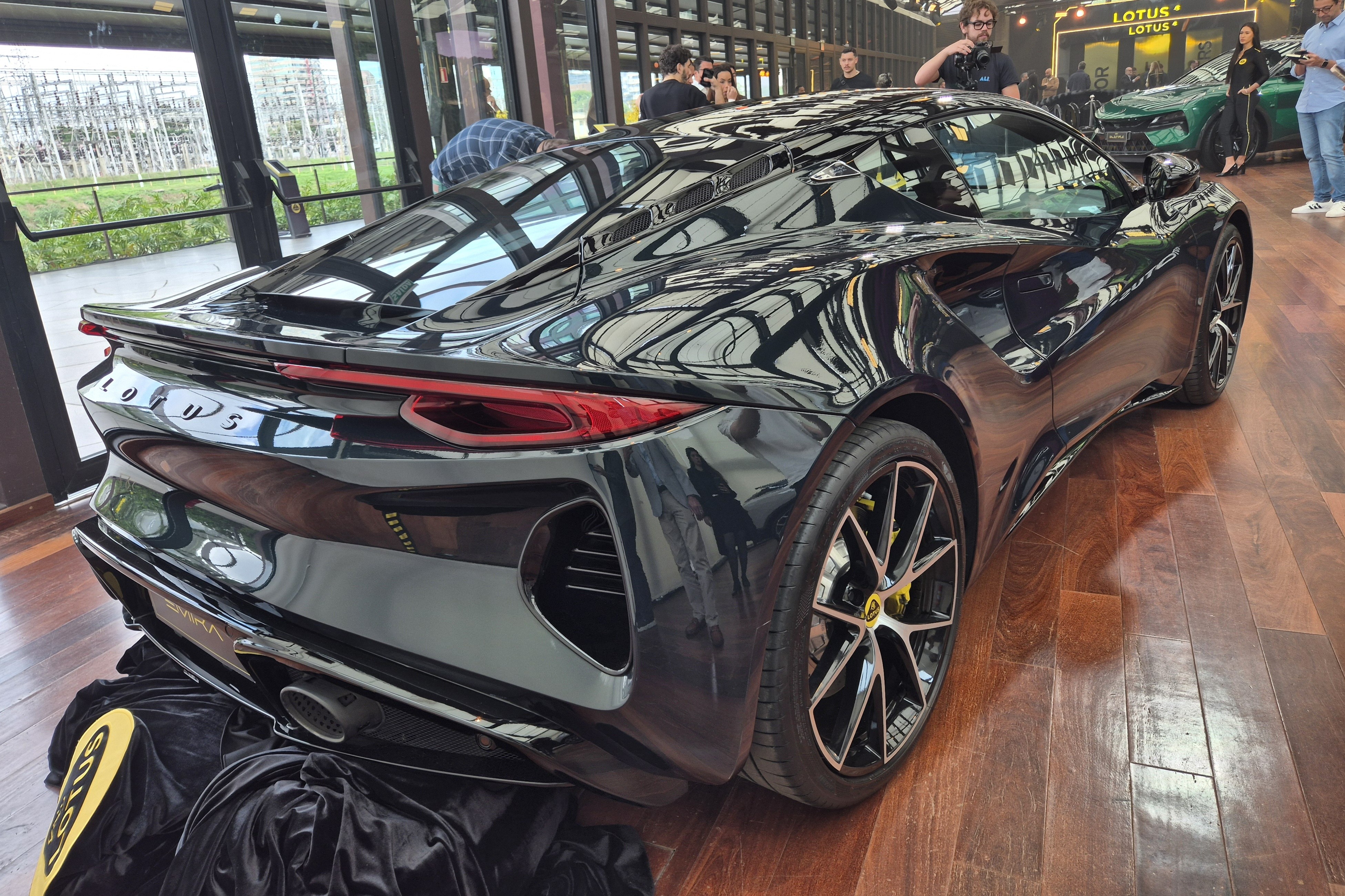 Carro esportivo Lotus Emira preto brilhante, visto de traseira e lateral, com lanternas vermelhas e rodas pretas com detalhes amarelos, exposto em piso de madeira. Pessoas observam ao fundo, e um SUV verde est&aacute; em destaque sob um letreiro 