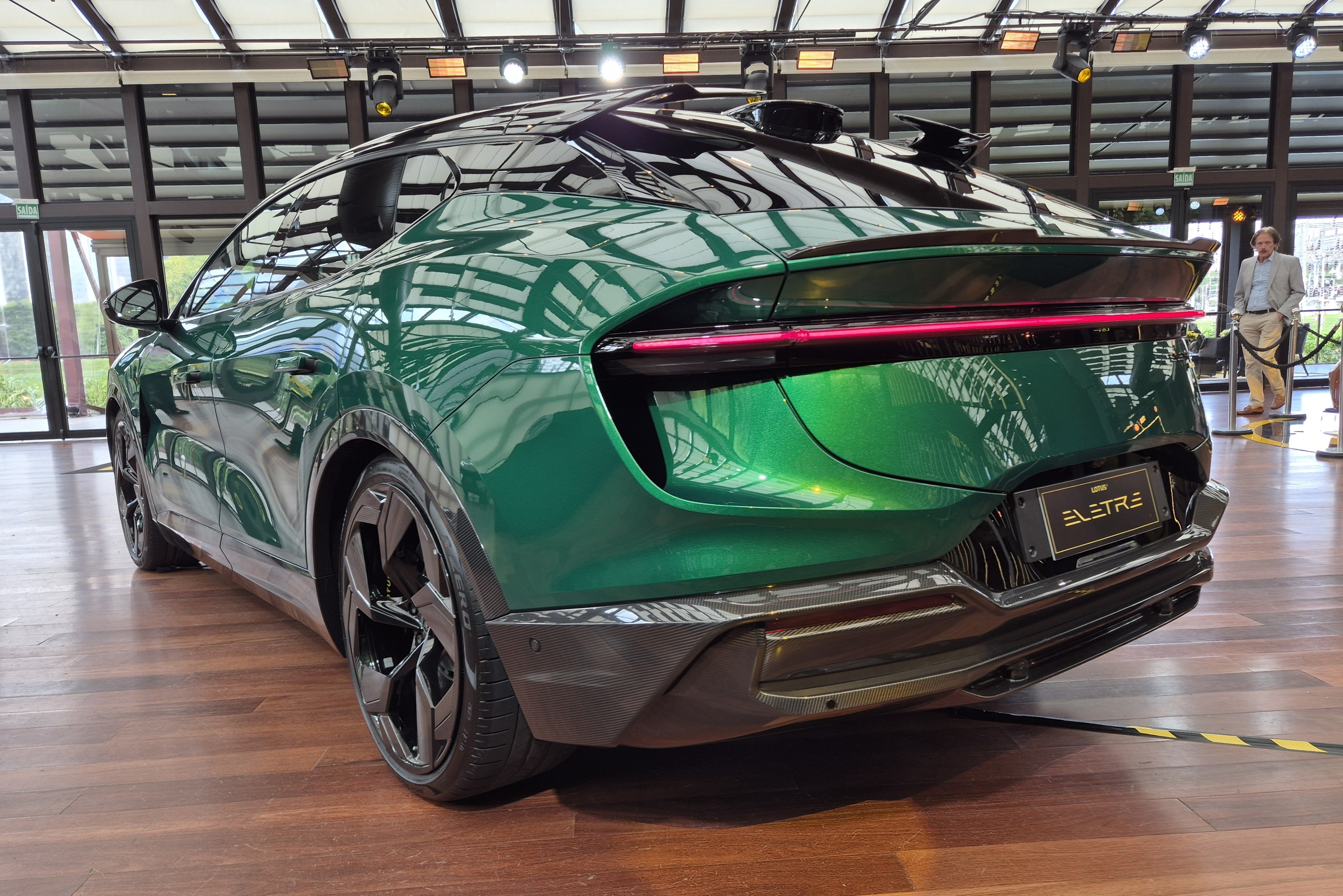 Traseira de um carro esportivo verde met&aacute;lico, modelo Lotus Eletre, com lanterna vermelha horizontal e aerof&oacute;lio duplo. O ve&iacute;culo est&aacute; em um piso de madeira, com um homem de blazer claro ao fundo, pr&oacute;ximo a uma janela.
