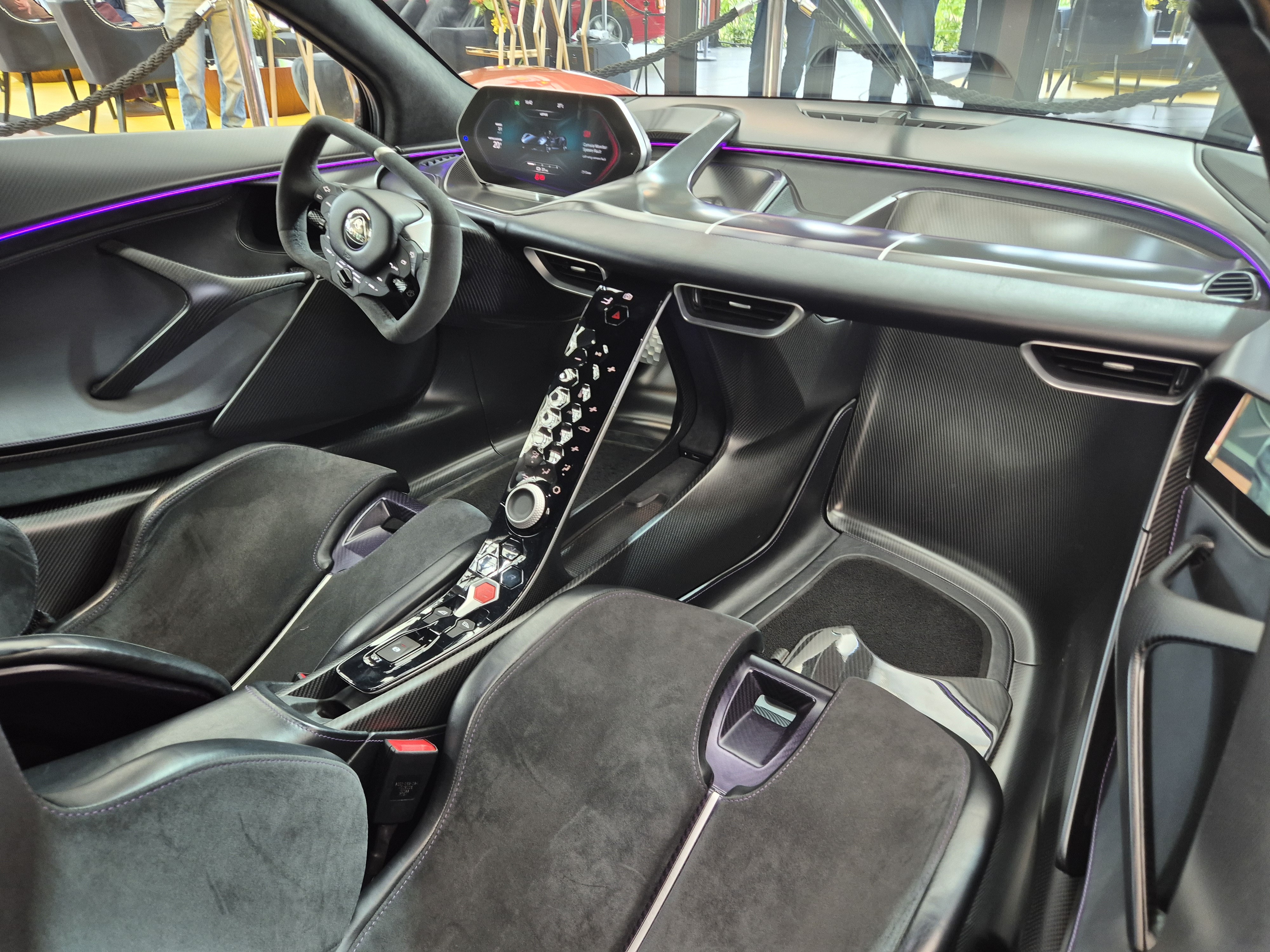 Interior de um carro esportivo moderno, com bancos de camur&ccedil;a preta, volante esportivo, painel digital e console central com bot&otilde;es futuristas. Uma linha de luz ambiente roxa percorre o painel, destacando o design arrojado.