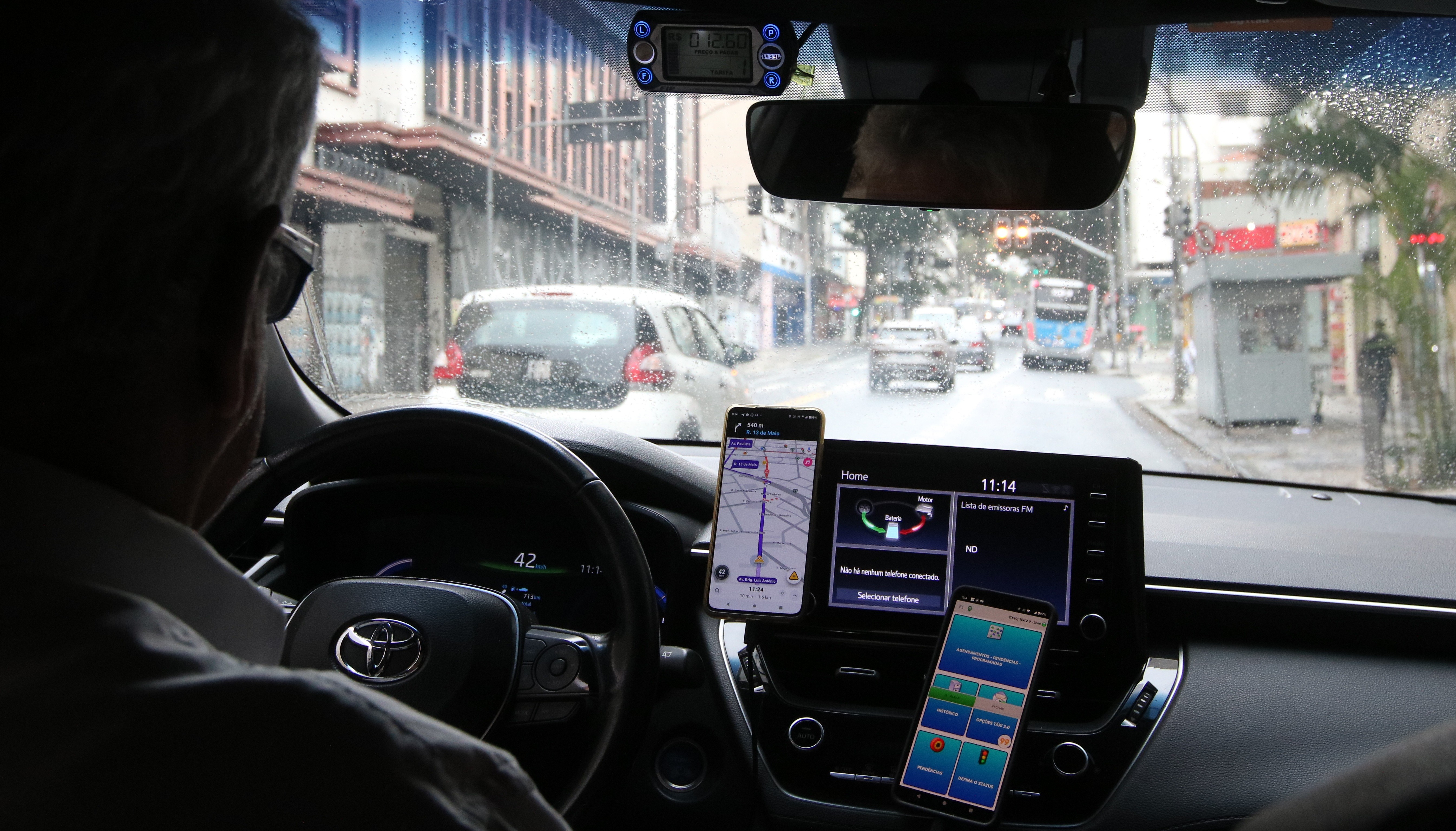Motorista de cabelos grisalhos ao volante de um carro, visto por tr&aacute;s, com tr&ecirc;s celulares e uma tela de navega&ccedil;&atilde;o no painel. O para-brisa molhado mostra uma rua movimentada com carros, um &ocirc;nibus e pedestres, sob um c&eacute;u nublado