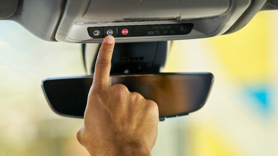Dedo indicador de uma pessoa pressionando o bot&atilde;o azul de um sistema de assist&ecirc;ncia veicular, localizado no console superior do carro, acima do retrovisor interno. H&aacute; tamb&eacute;m um bot&atilde;o vermelho de SOS e outro com um &iacute;cone de telefone