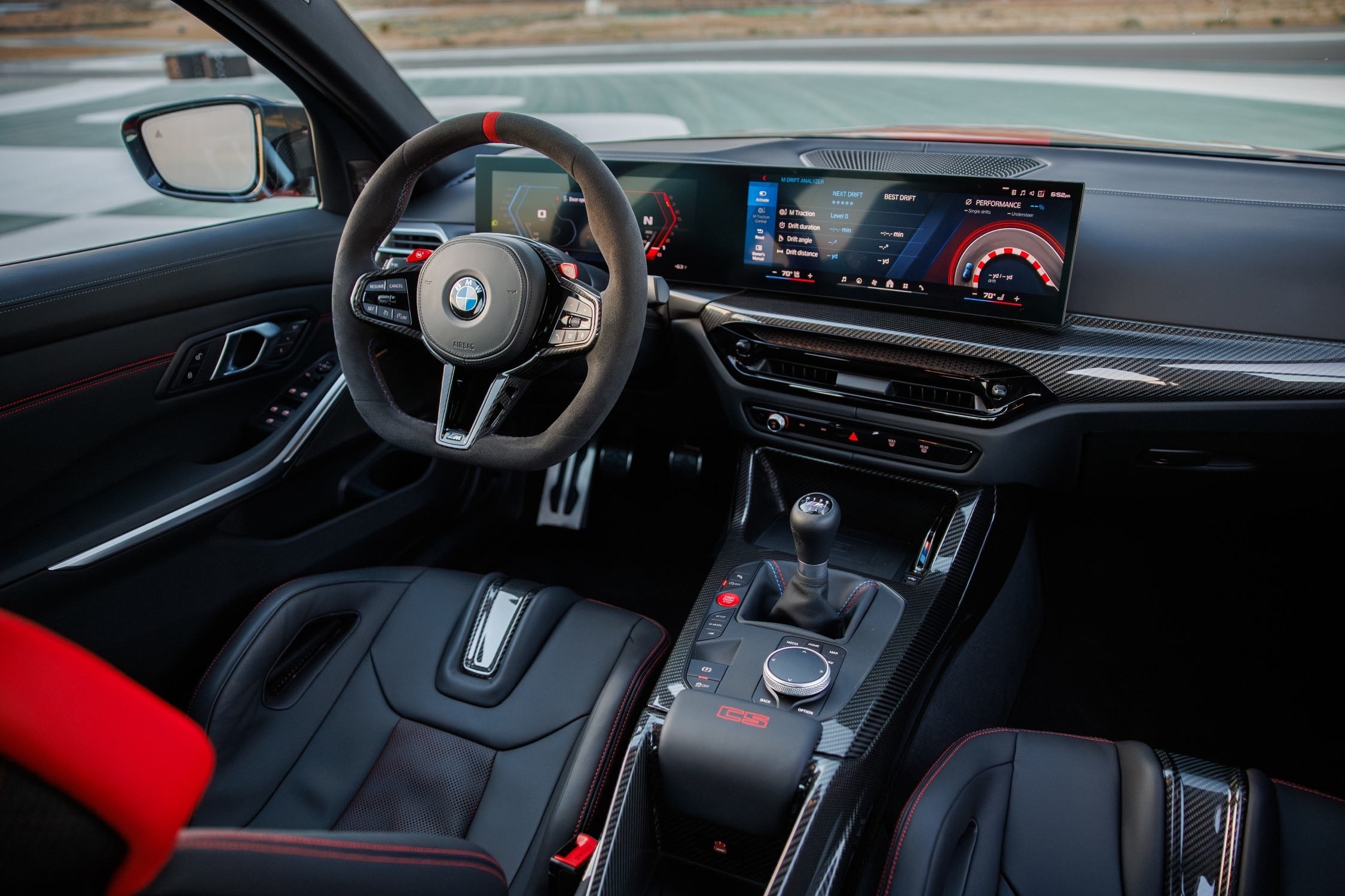 Interior de um carro esportivo BMW, com volante de camur&ccedil;a e detalhe vermelho, painel digital exibindo informa&ccedil;&otilde;es de desempenho e um mapa de pista, c&acirc;mbio manual, bancos de couro preto com costura vermelha e detalhes em fibra de carbono no painel e console central