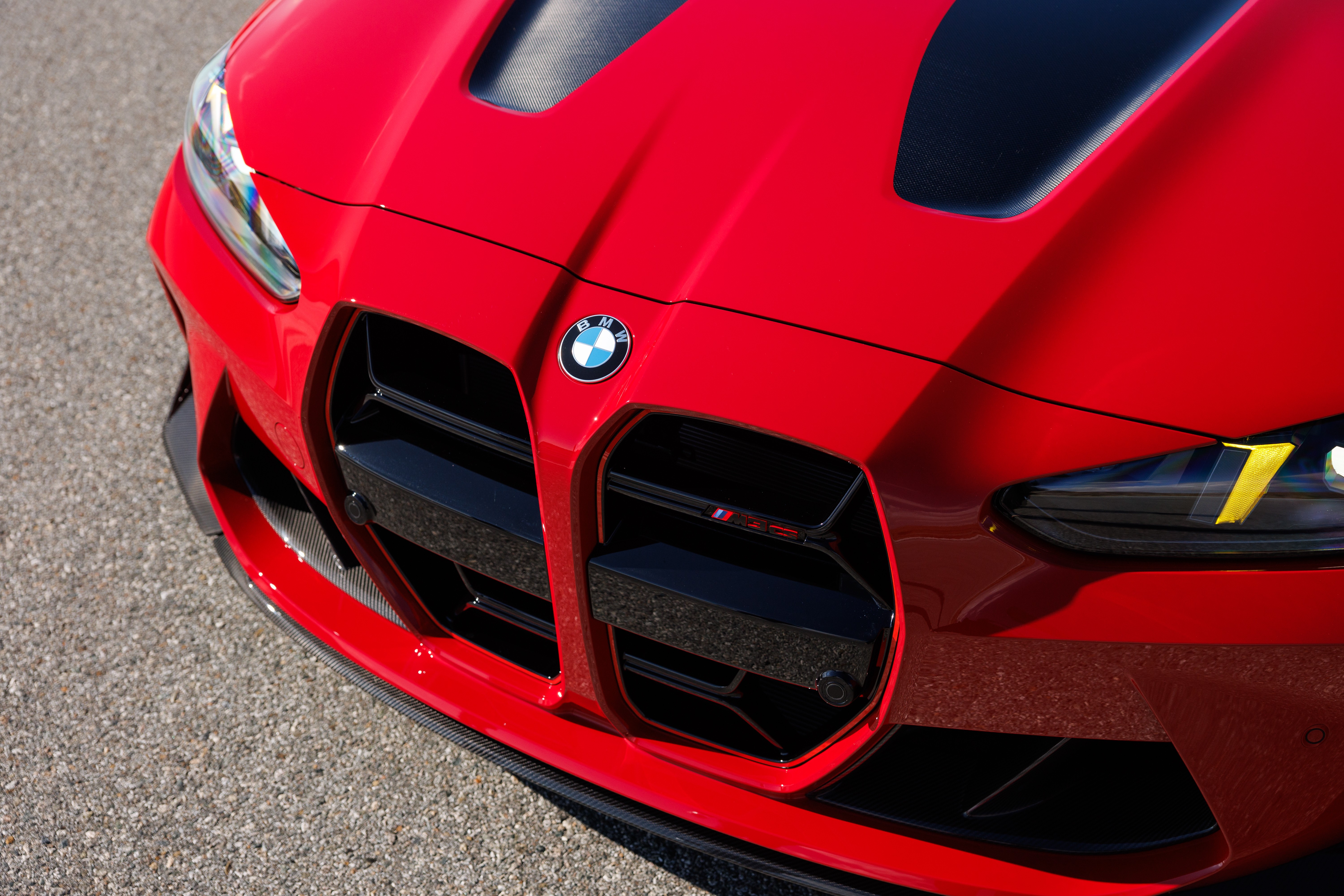 Frente de um carro esportivo BMW vermelho, com grade dupla preta, far&oacute;is de LED e detalhes em fibra de carbono no cap&ocirc; e para-choque, estacionado em asfalto cinza