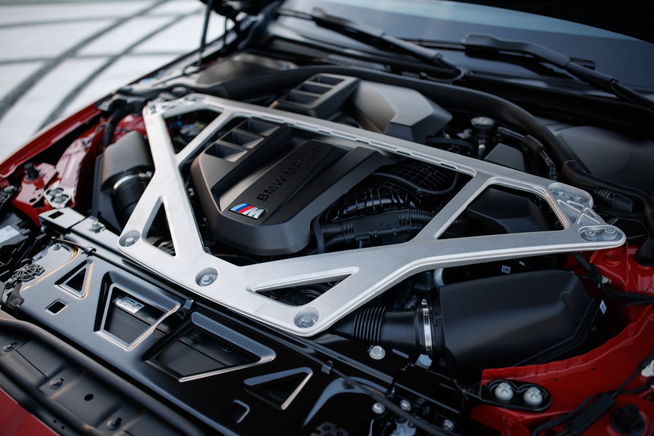 Motor de carro esportivo BMW M com tampa preta e logo M colorido, cercado por componentes prateados e vermelhos, sob o cap&ocirc; aberto