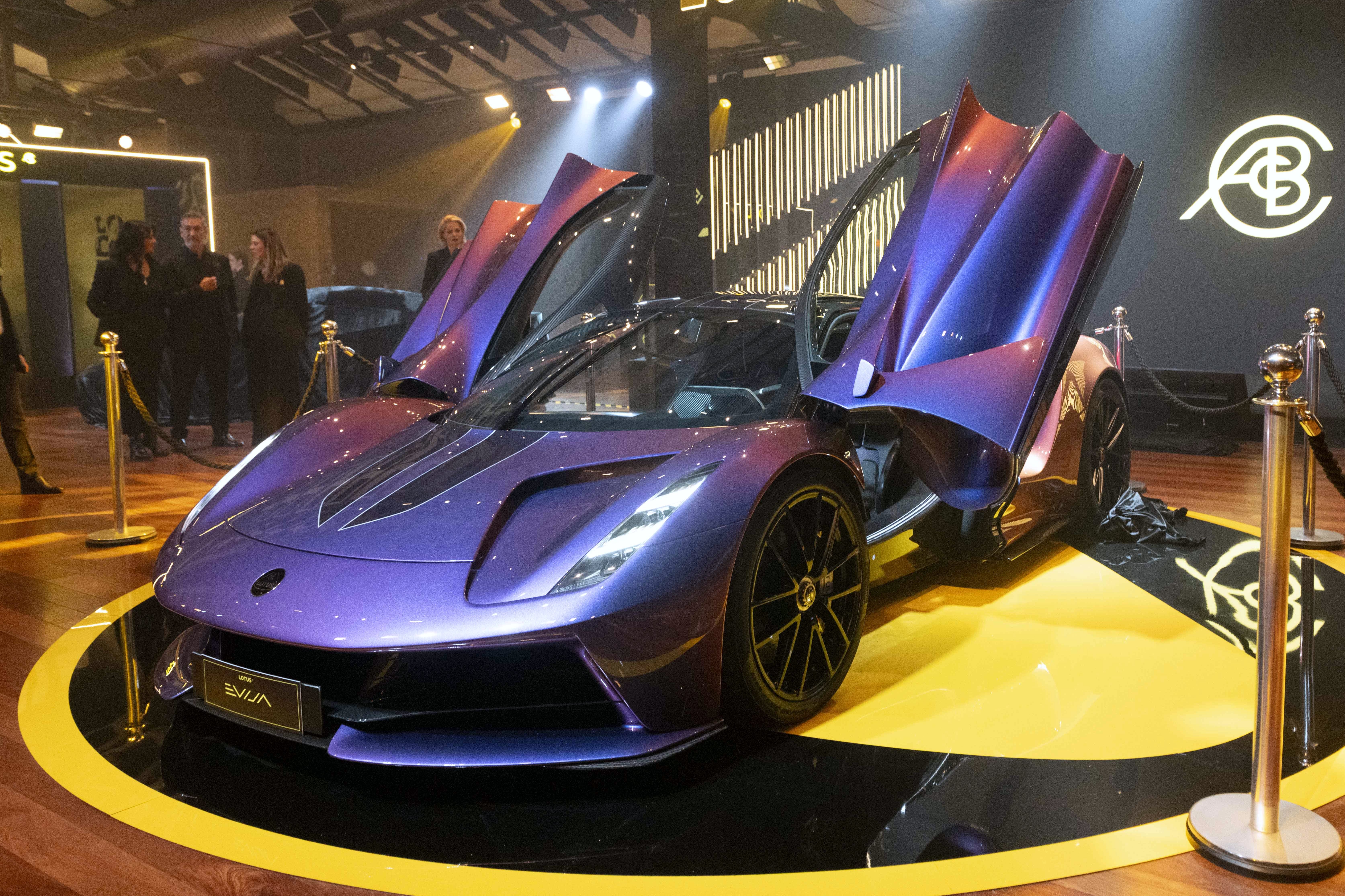 Carro esportivo roxo met&aacute;lico com portas abertas para cima, exibido em plataforma preta e amarela. Pessoas observam ao fundo, em ambiente com ilumina&ccedil;&atilde;o focada e um logotipo branco na parede