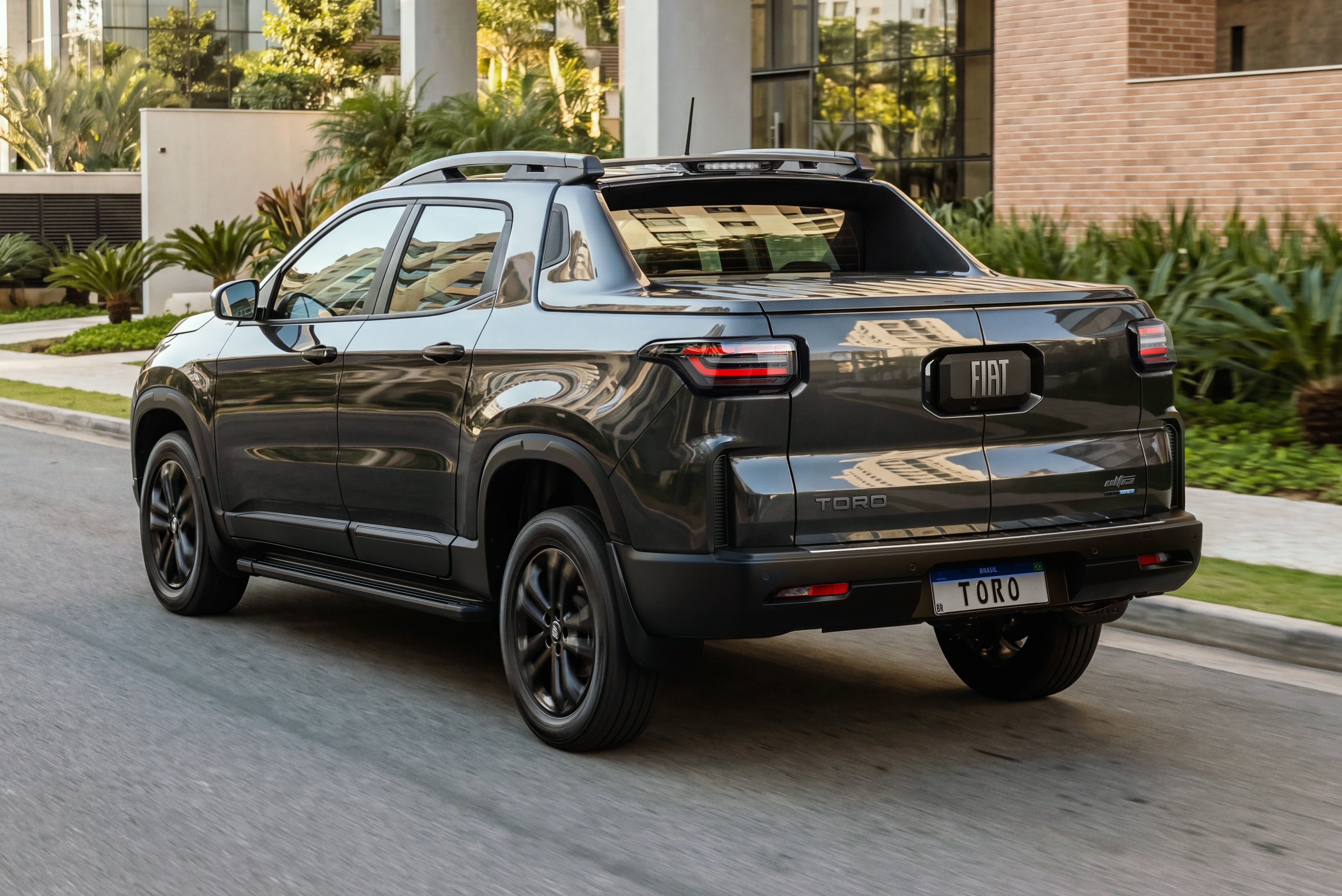 Picape Fiat Toro cinza escura vista de traseira e lateral, com rodas pretas, em rua asfaltada. Ao fundo, pr&eacute;dios modernos e vegeta&ccedil;&atilde;o exuberante