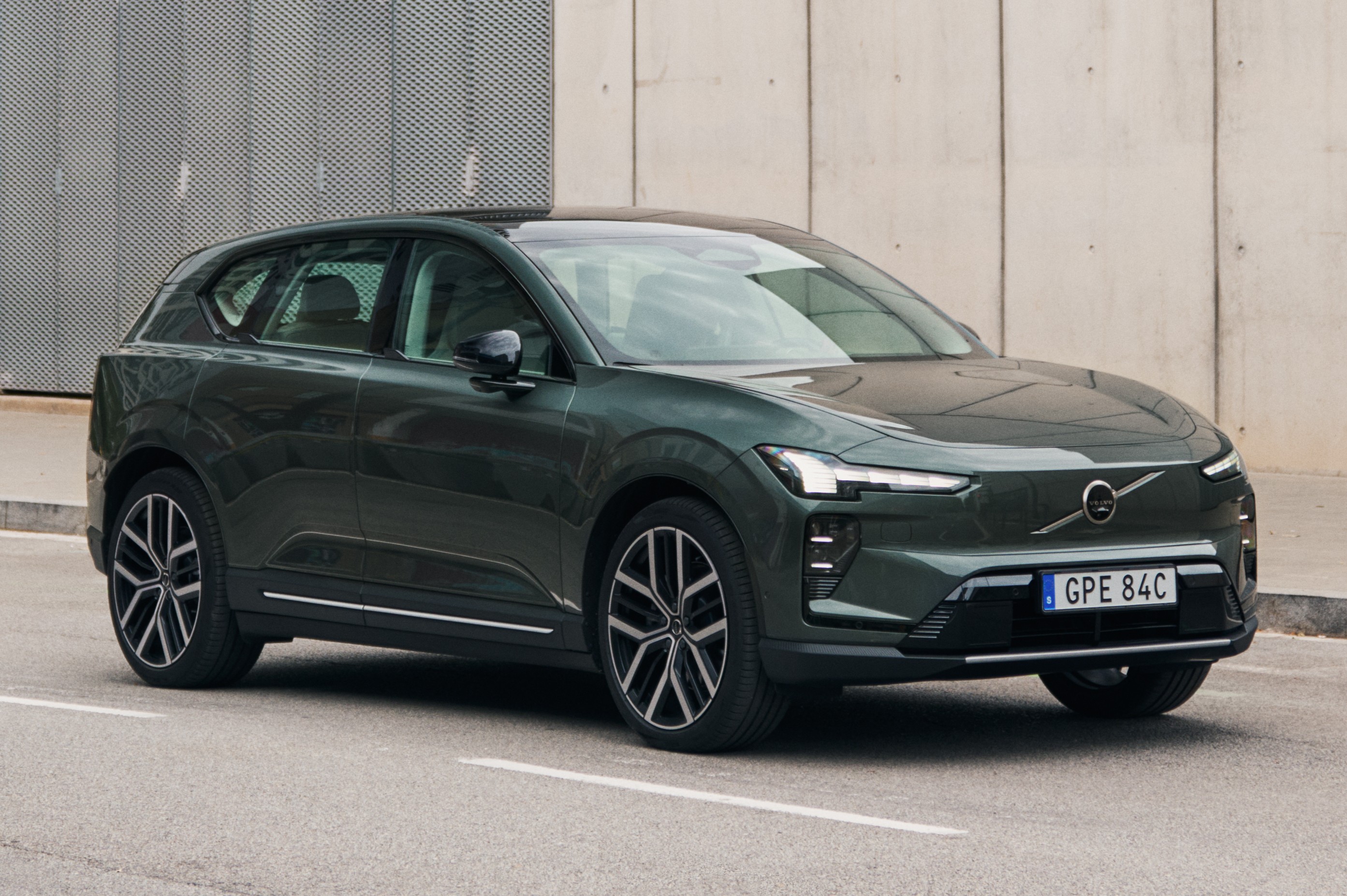 SUV el&eacute;trico Volvo EX30 verde escuro, visto de frente e lateral, parado em uma rua asfaltada com faixas brancas, em frente a uma parede cinza texturizada