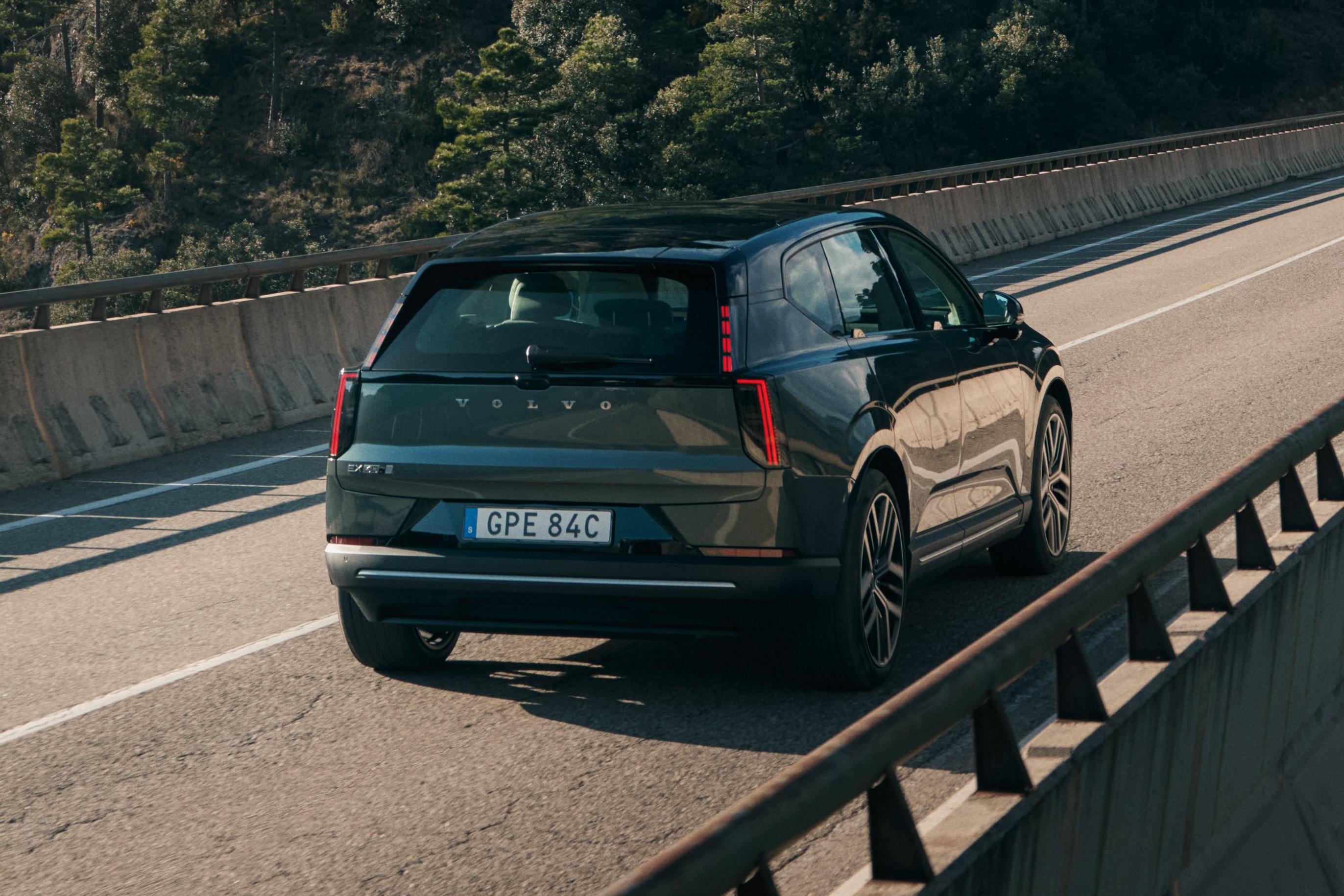 SUV Volvo EX90 escuro em uma estrada de asfalto, visto por tr&aacute;s, com montanhas arborizadas ao fundo