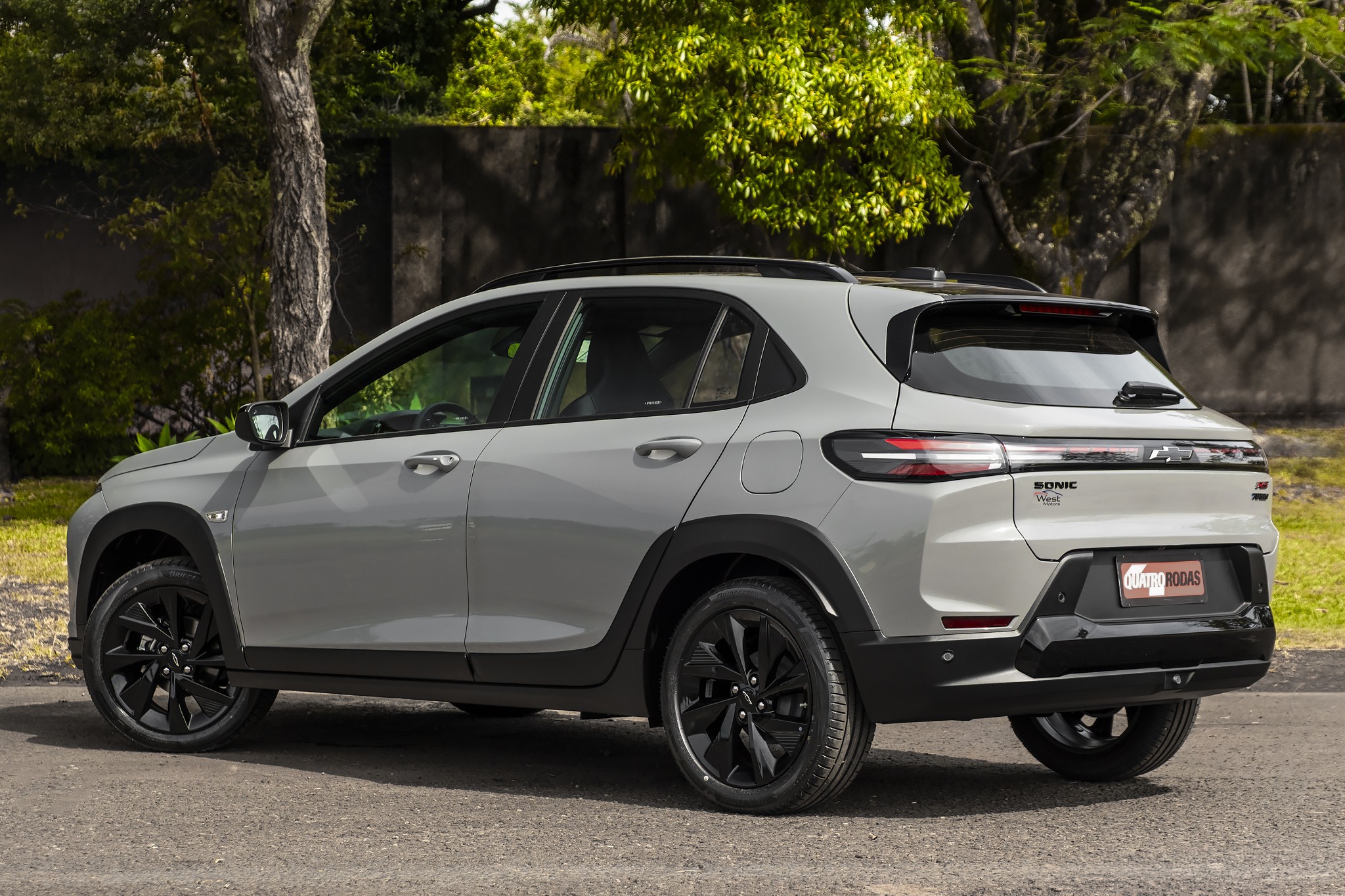 SUV compacto cinza claro, com rodas e detalhes em preto, estacionado em uma rua de terra com &aacute;rvores e vegeta&ccedil;&atilde;o ao fundo