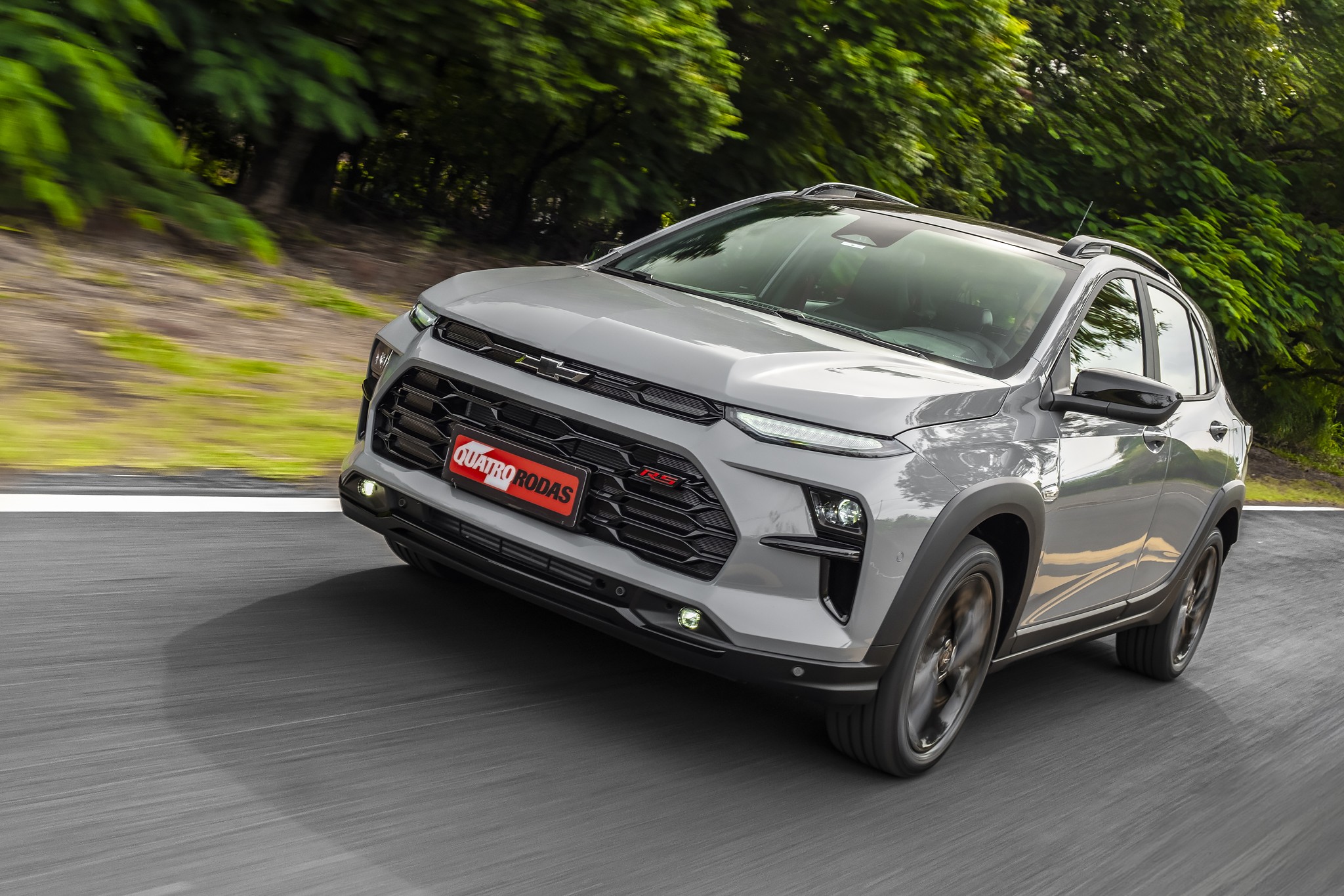 SUV Chevrolet Sonic cinza claro em movimento, com a frente virada para a esquerda, em uma estrada asfaltada com &aacute;rvores verdes ao fundo
