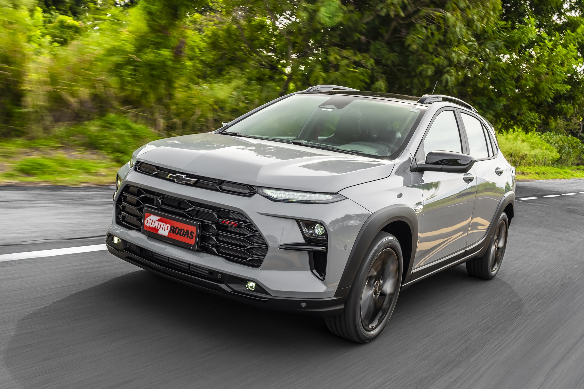SUV Chevrolet Sonic cinza claro em movimento numa estrada asfaltada, com vegeta&ccedil;&atilde;o verde borrada ao fundo