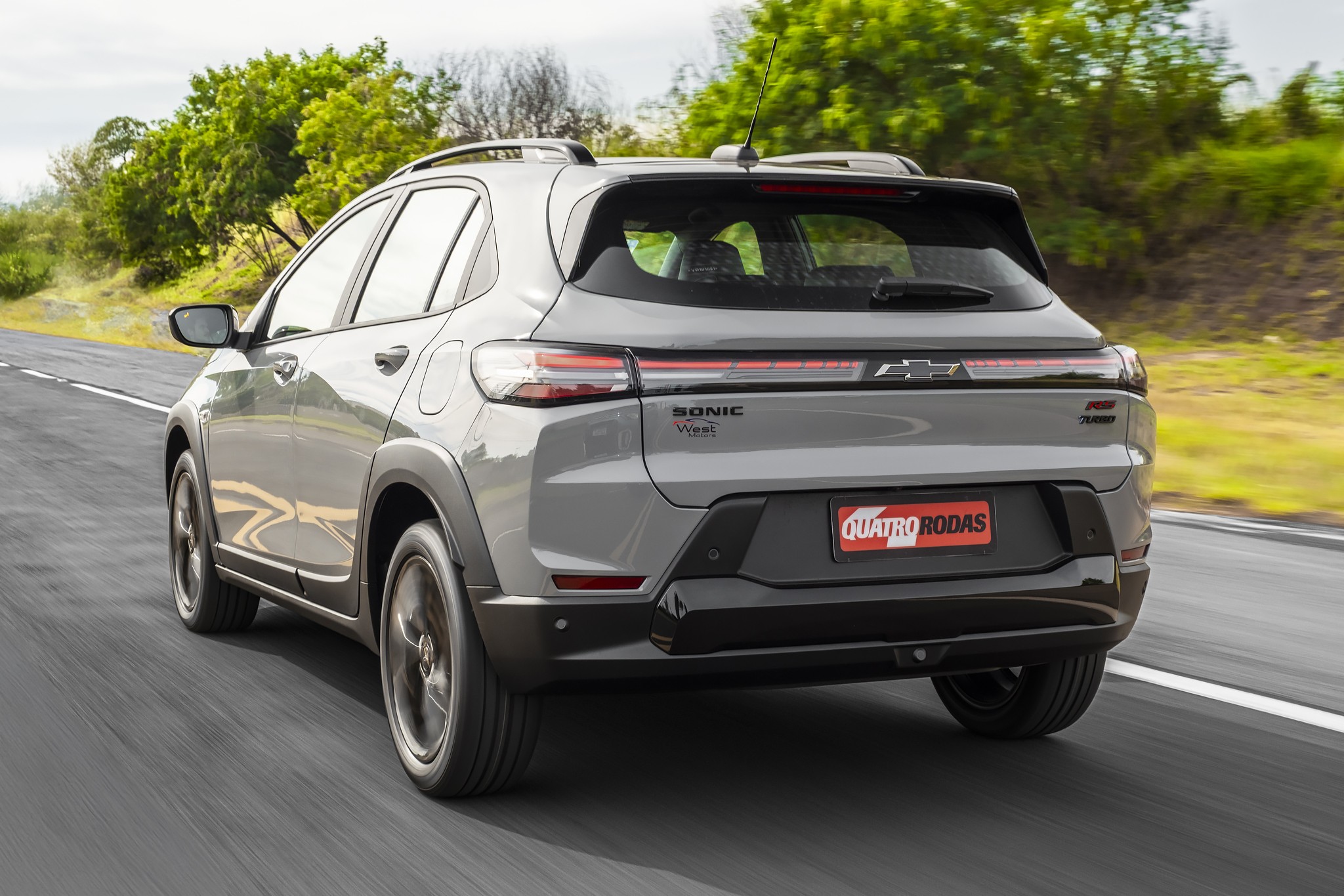 SUV Chevrolet Sonic cinza escuro visto por tr&aacute;s, em movimento numa estrada com vegeta&ccedil;&atilde;o ao fundo
