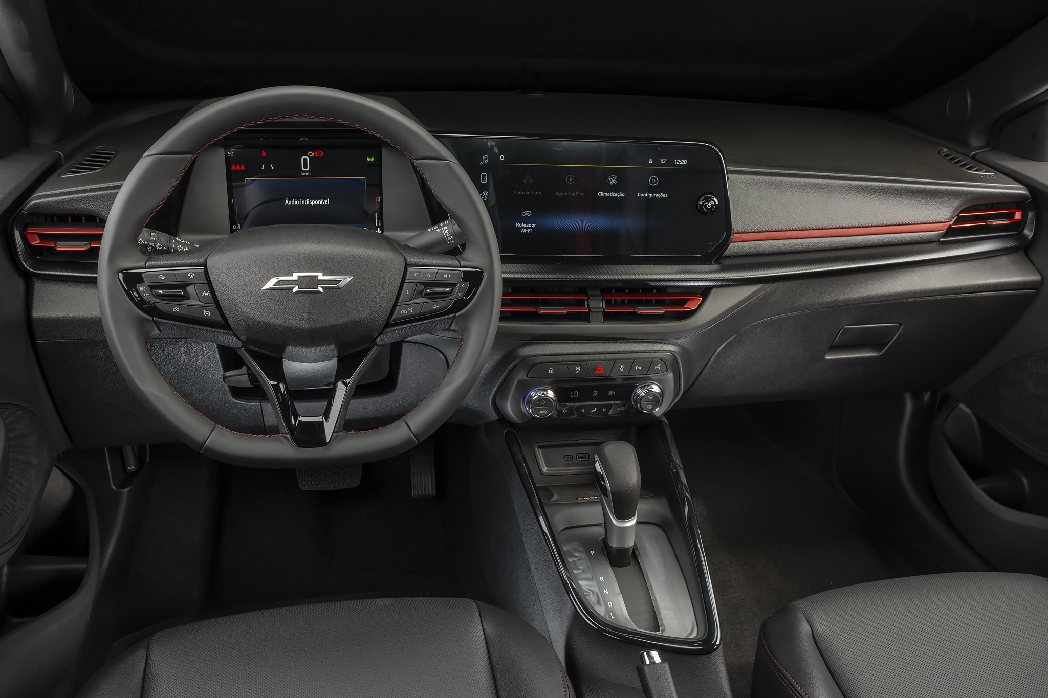 Interior de carro moderno com painel escuro, volante multifuncional com logo Chevrolet, tela digital do painel e central multim&iacute;dia. Detalhes em vermelho nas sa&iacute;das de ar e friso horizontal. C&acirc;mbio autom&aacute;tico e bancos de couro pretos vis&iacute;veis