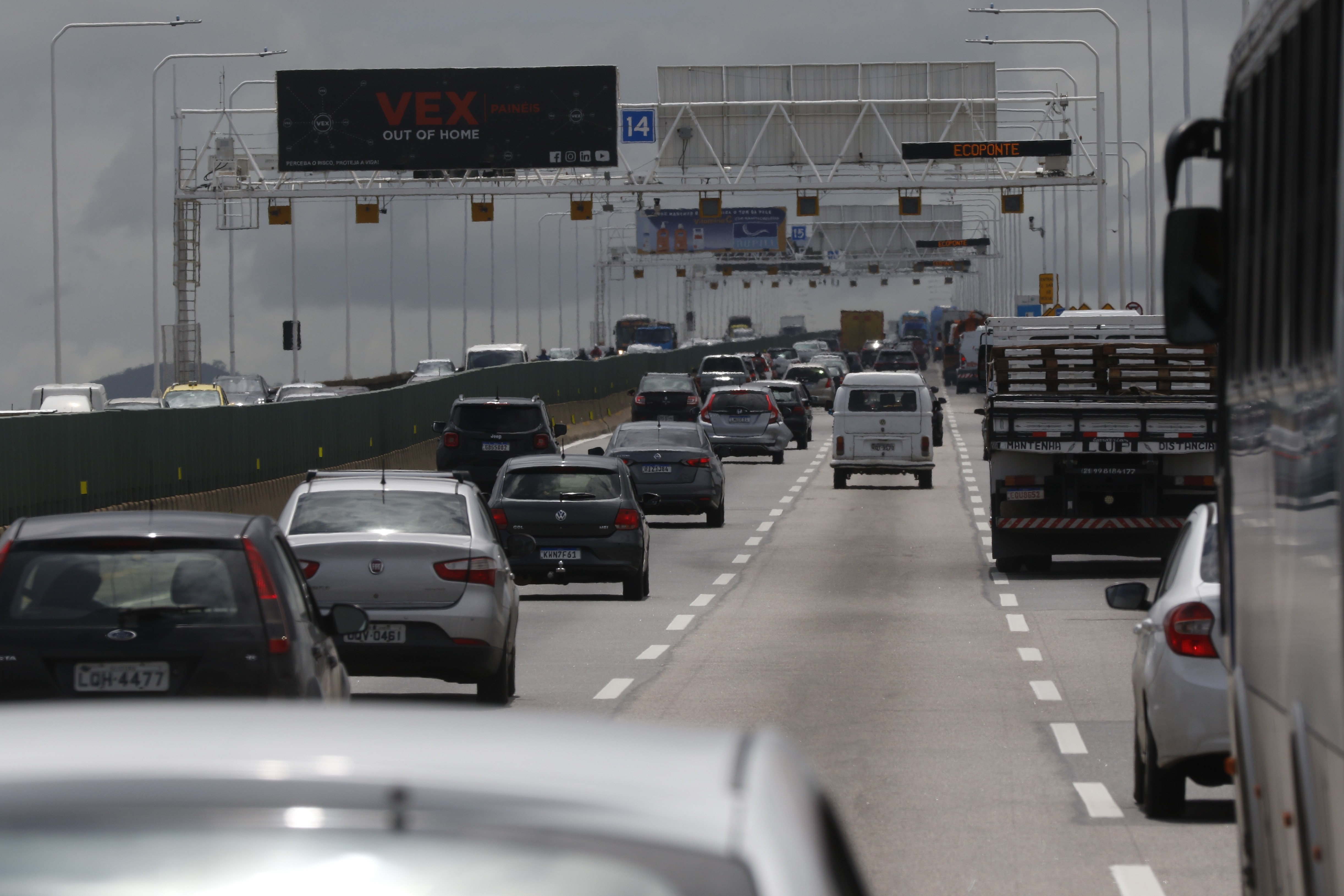 Tr&aacute;fego intenso em uma rodovia com v&aacute;rios carros, caminh&otilde;es e uma van branca. No fundo, uma placa eletr&ocirc;nica preta com a palavra 