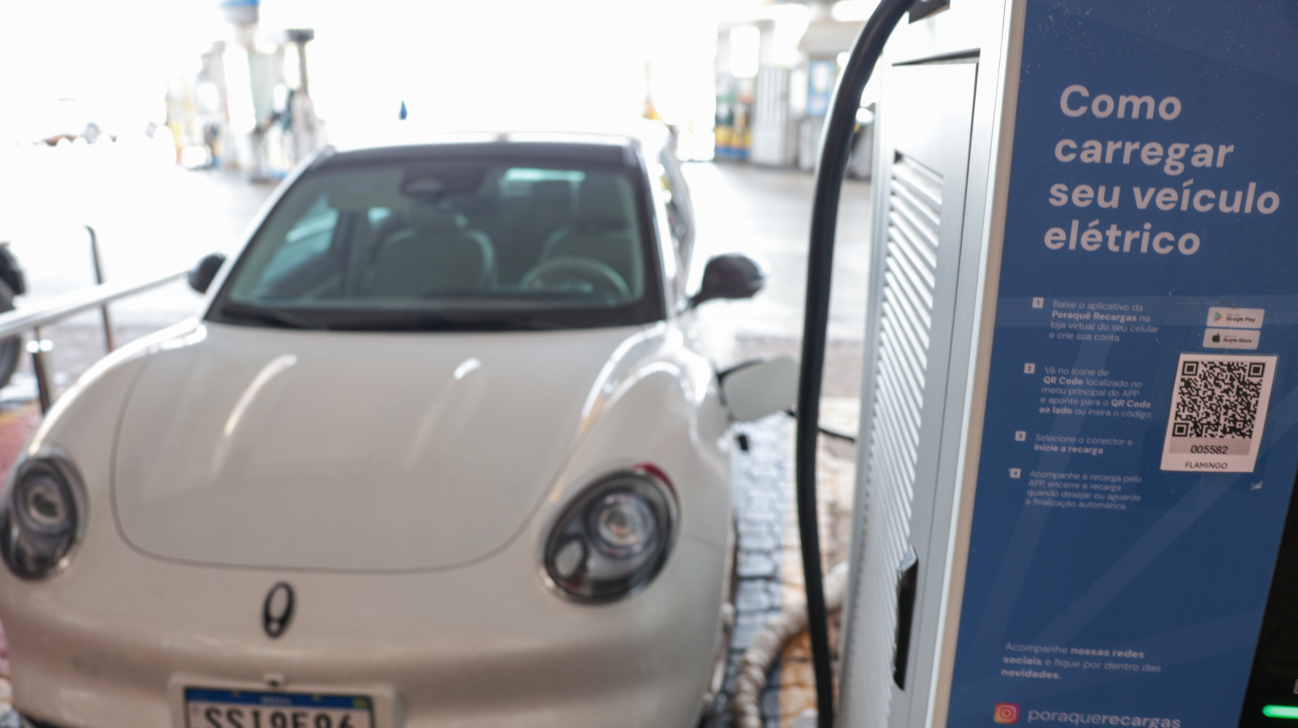 Carro el&eacute;trico branco, modelo compacto, sendo carregado em um posto com painel azul que exibe 