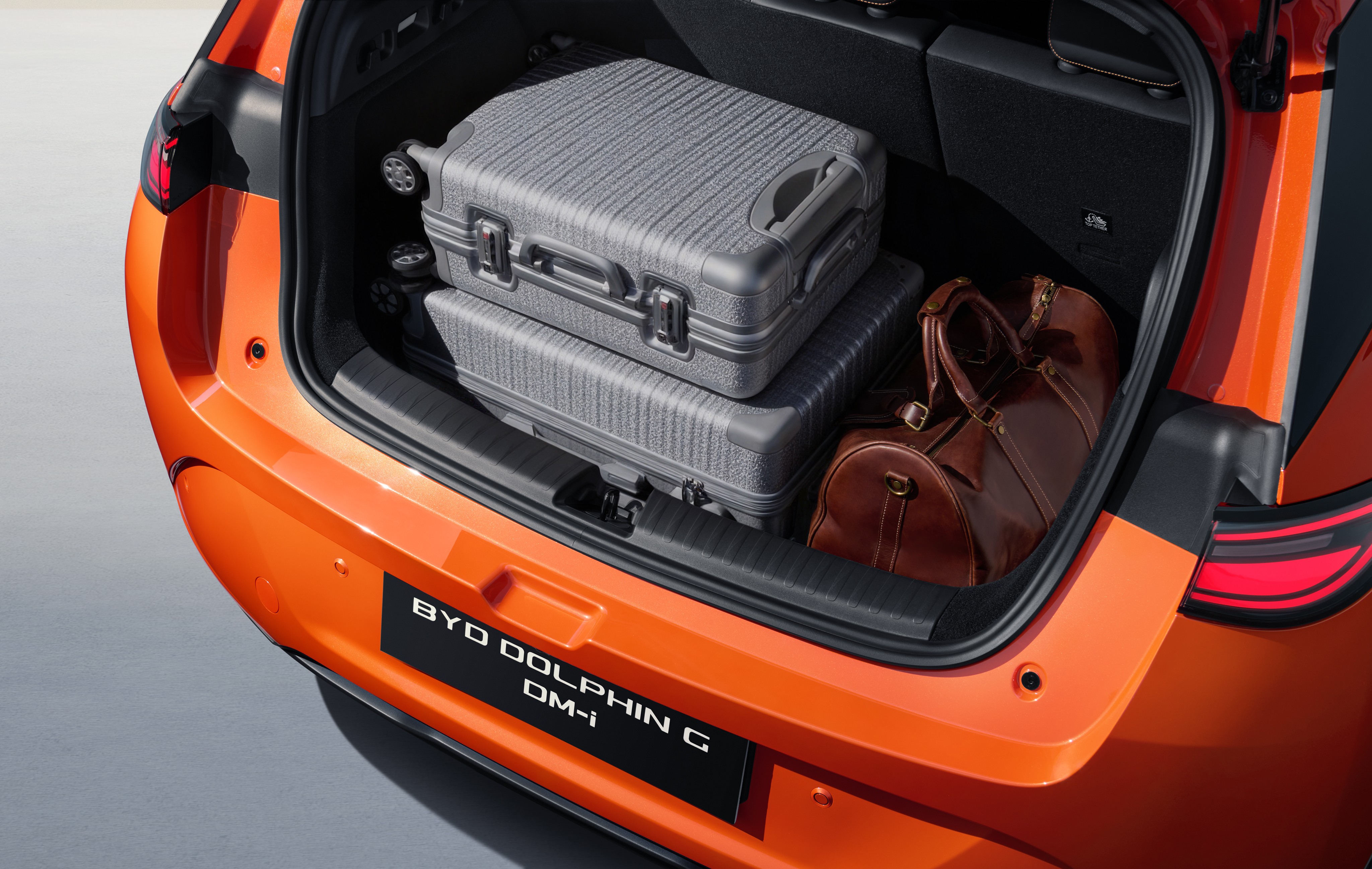 Porta-malas de um carro laranja aberto, com duas malas cinzas empilhadas e uma bolsa de viagem marrom ao lado. A placa preta do carro exibe BYD DOLPHIN G DM-i em branco