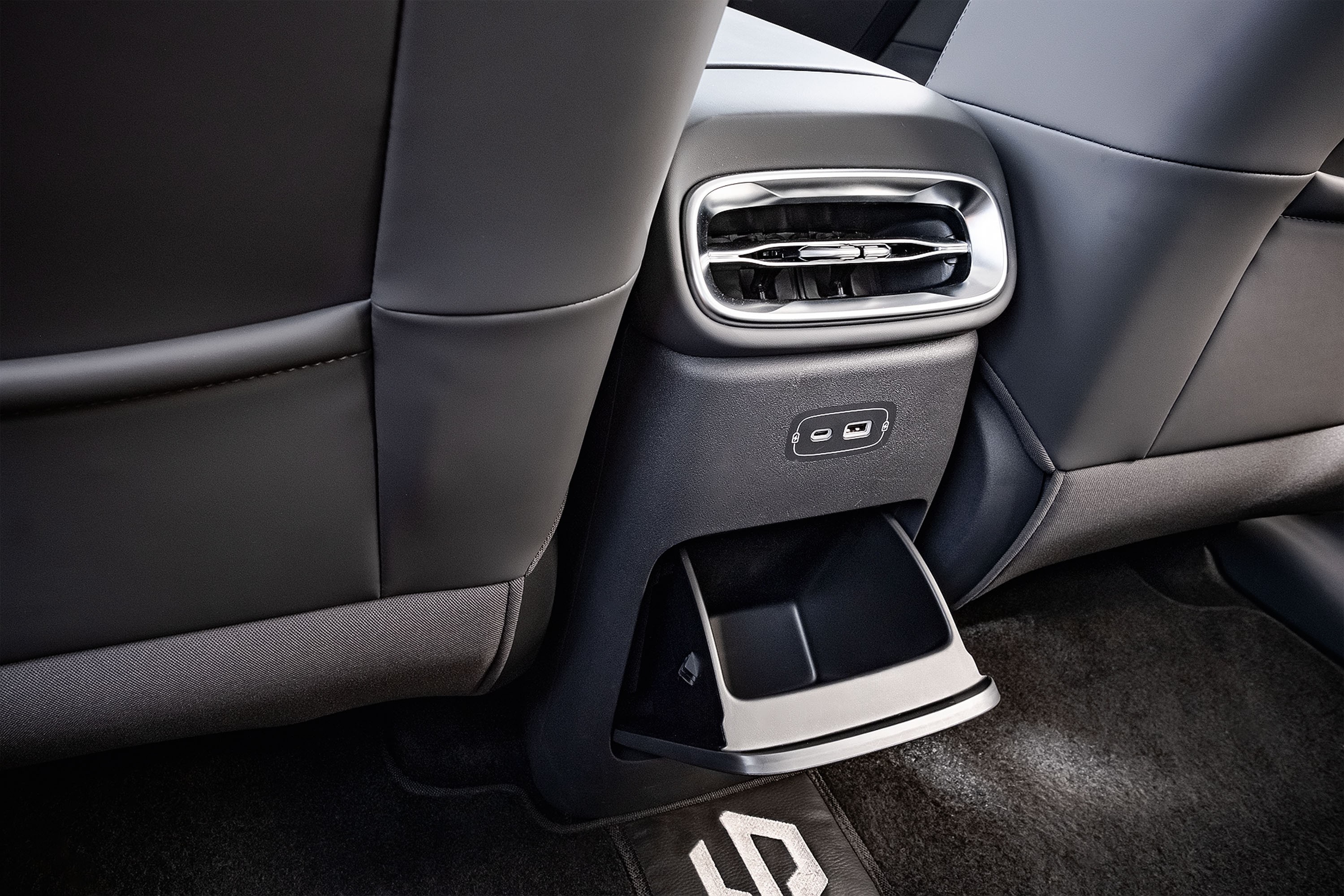 Interior de carro com bancos traseiros de couro preto, console central com sa&iacute;das de ar cromadas, duas portas USB-C e um porta-objetos retr&aacute;til, sobre tapete escuro com logo