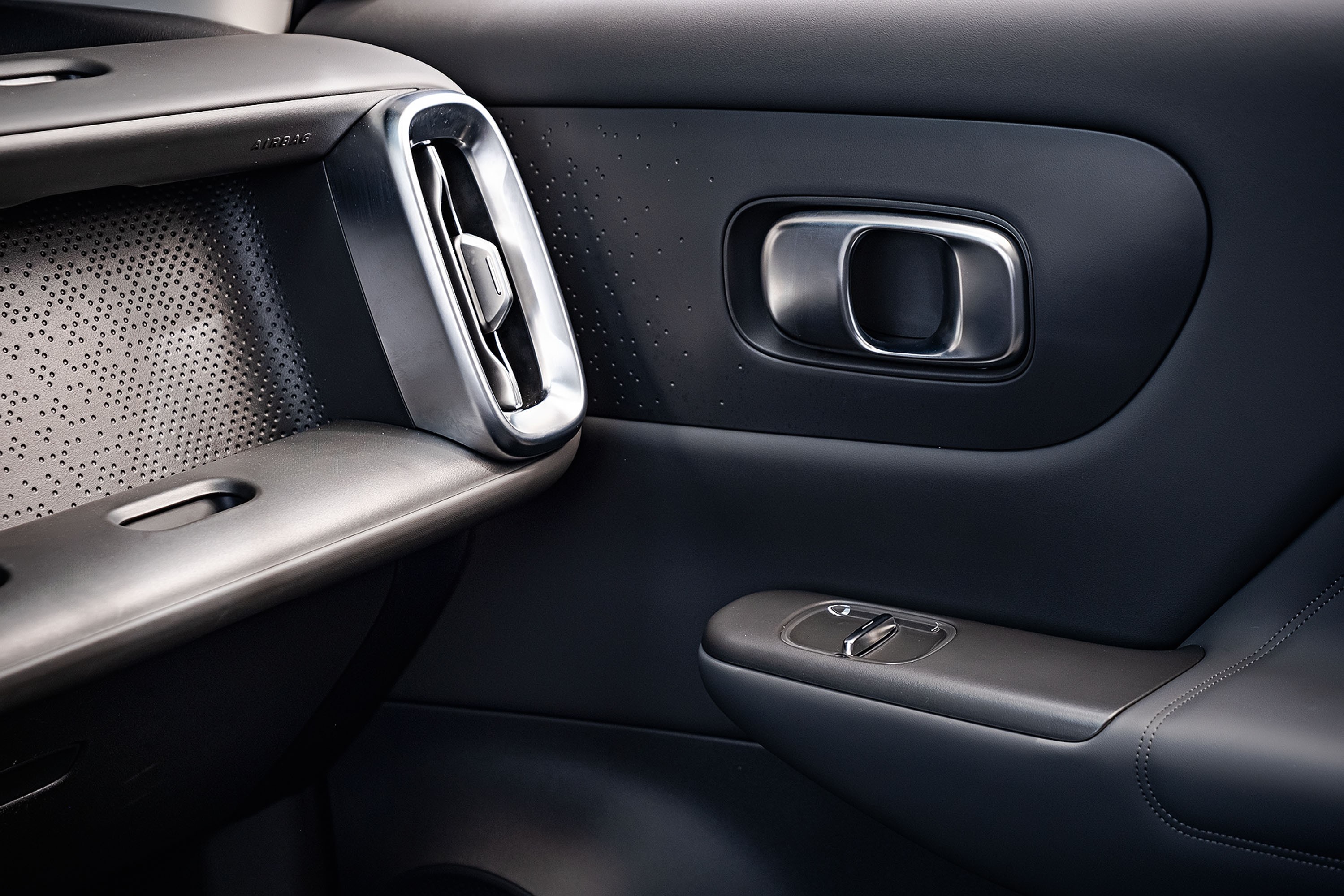 Interior de carro moderno com detalhes em cinza escuro e prata. &Agrave; esquerda, painel texturizado com pequenos furos e sa&iacute;da de ar vertical cromada. &Agrave; direita, ma&ccedil;aneta da porta em metal escovado e, abaixo, controles dos vidros el&eacute;tricos com acabamento em cinza e prata