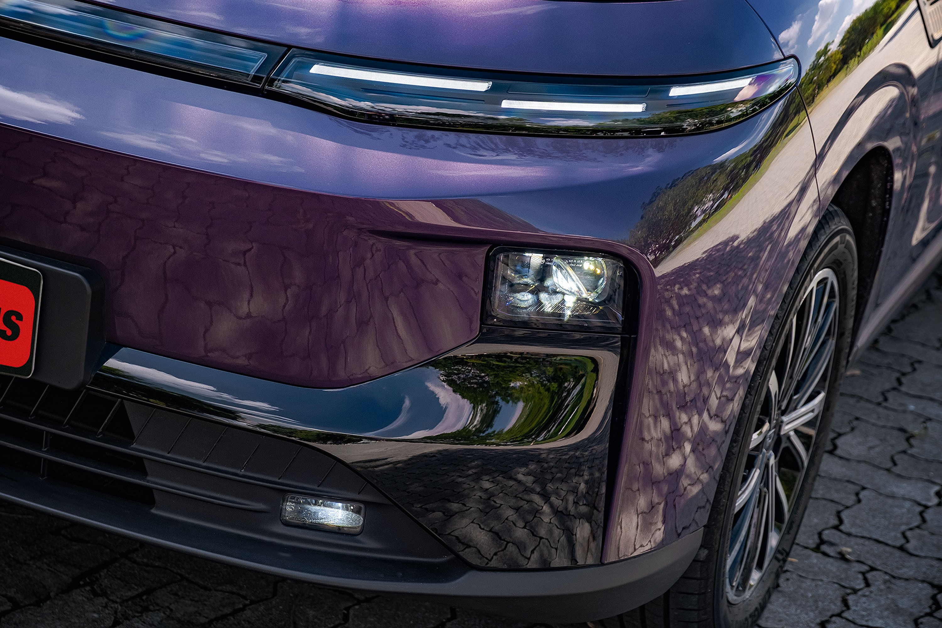 Parte frontal de um carro roxo met&aacute;lico, com far&oacute;is de LED horizontais e um farol principal quadrado aceso. A superf&iacute;cie reflete o c&eacute;u e &aacute;rvores, e a roda com aros prateados &eacute; vis&iacute;vel &agrave; direita. O ve&iacute;culo est&aacute; sobre um piso de paralelep&iacute;pedos