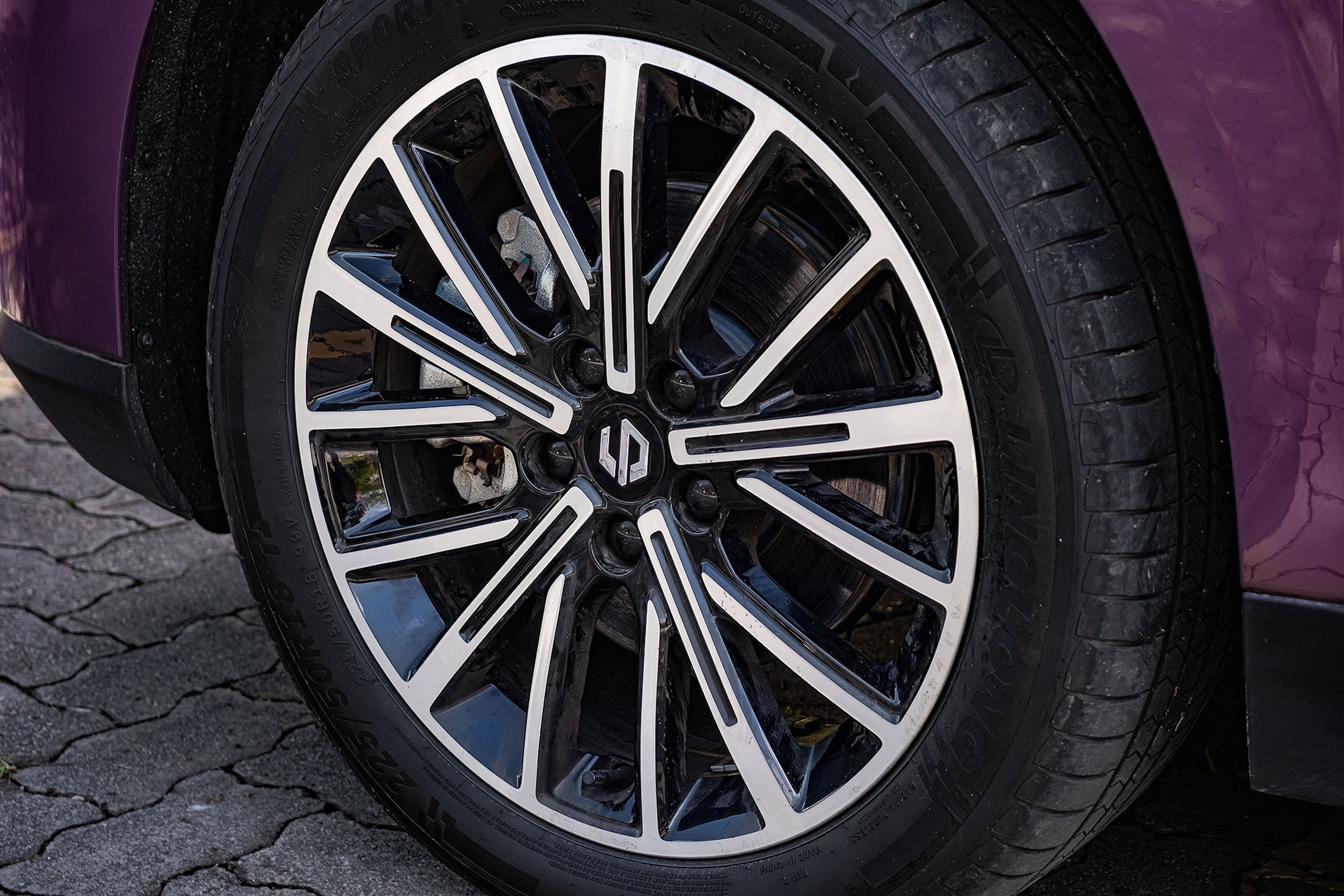 Roda de carro com aro preto e detalhes prateados, pneu com sulcos vis&iacute;veis, parte da lataria roxa e ch&atilde;o de paralelep&iacute;pedos
