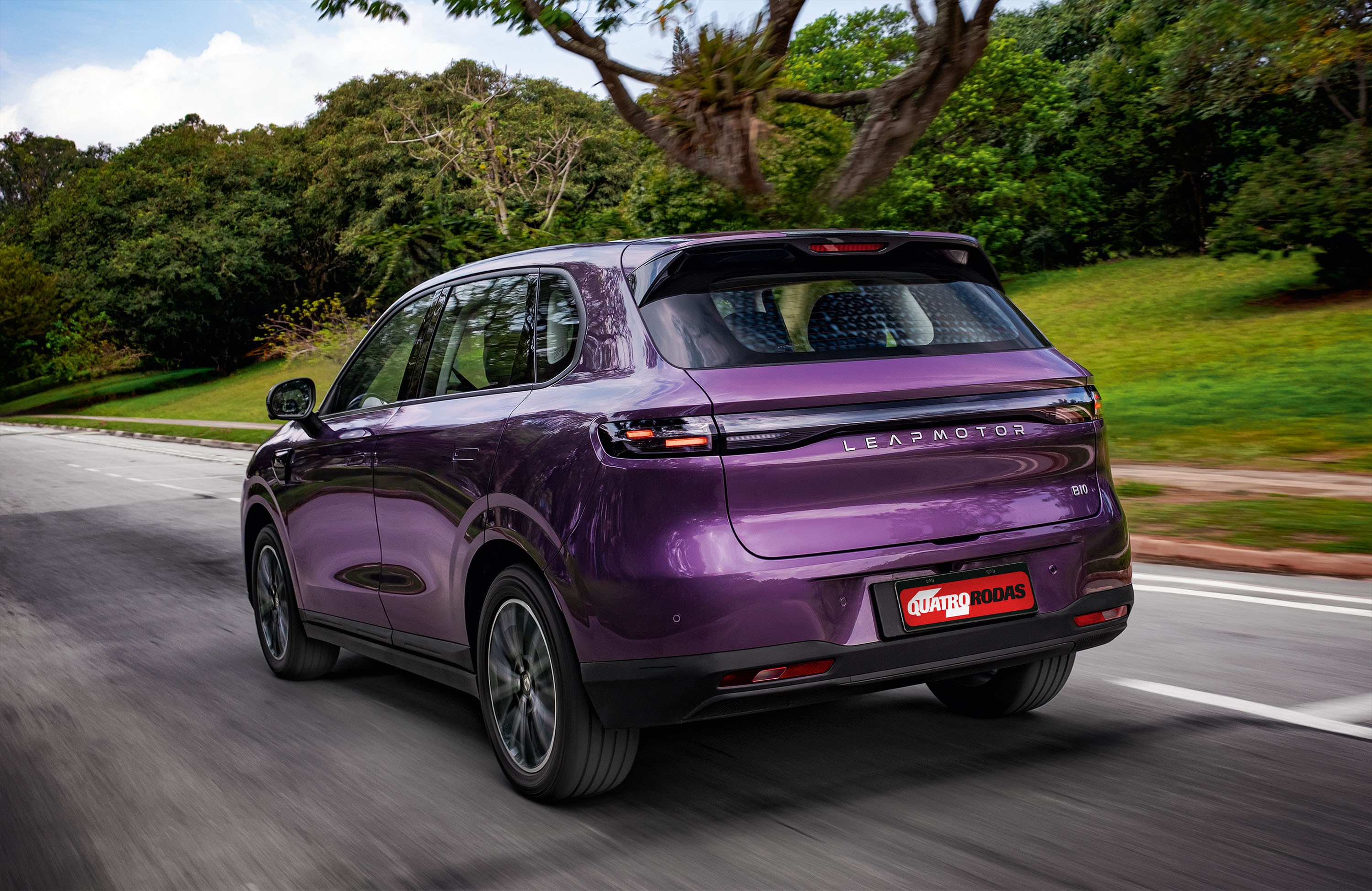 SUV Leapmotor C10 roxo met&aacute;lico em movimento, visto por tr&aacute;s, em uma estrada asfaltada com &aacute;rvores verdes ao fundo e c&eacute;u azul com nuvens