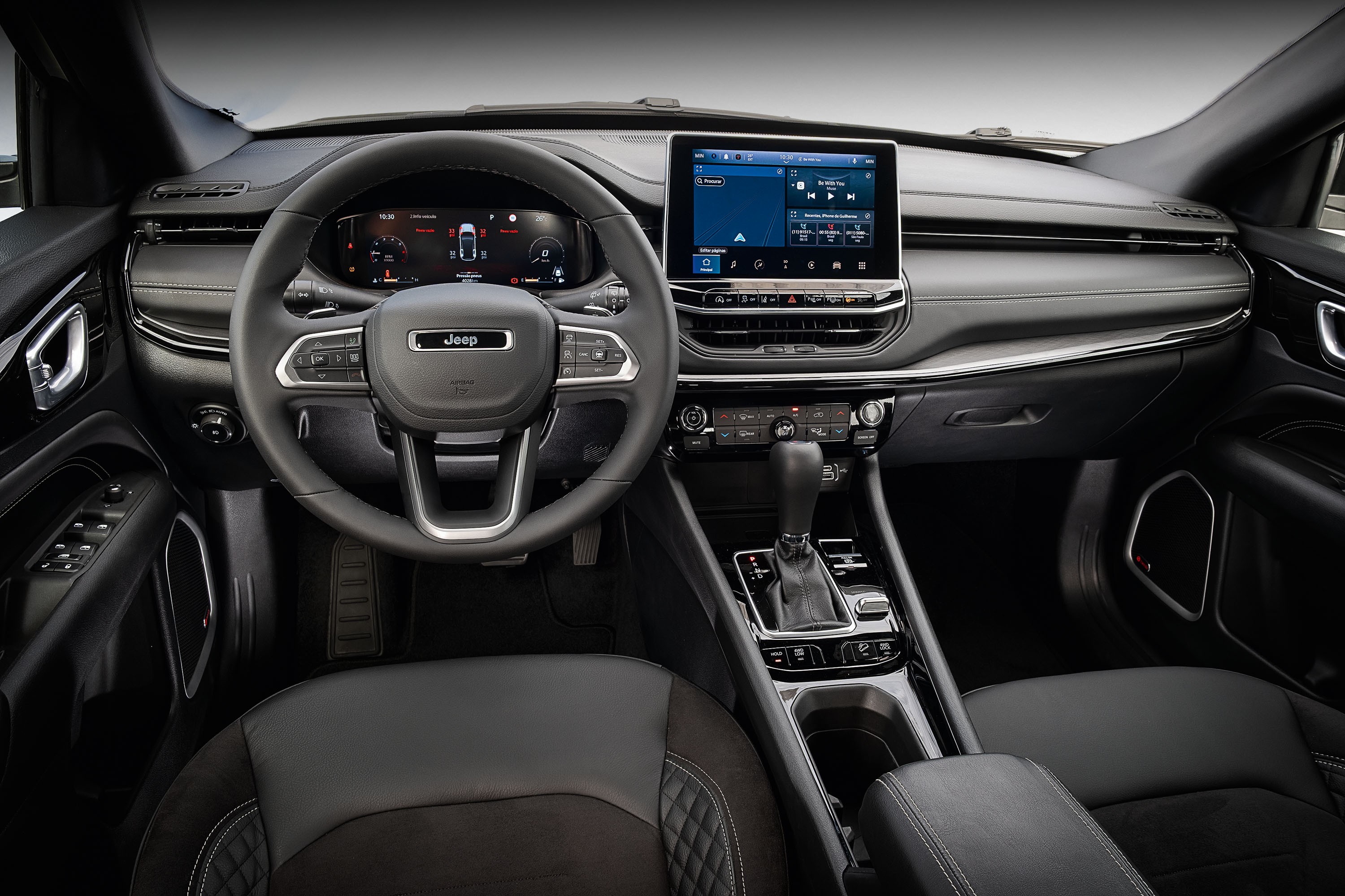 Interior de um carro moderno em tons escuros, com volante multifuncional da marca Jeep, painel digital e tela multim&iacute;dia central exibindo um reprodutor de m&uacute;sica. O console central possui c&acirc;mbio autom&aacute;tico e bot&otilde;es de controle, e o banco do motorista &eacute; de couro preto com costura em losango