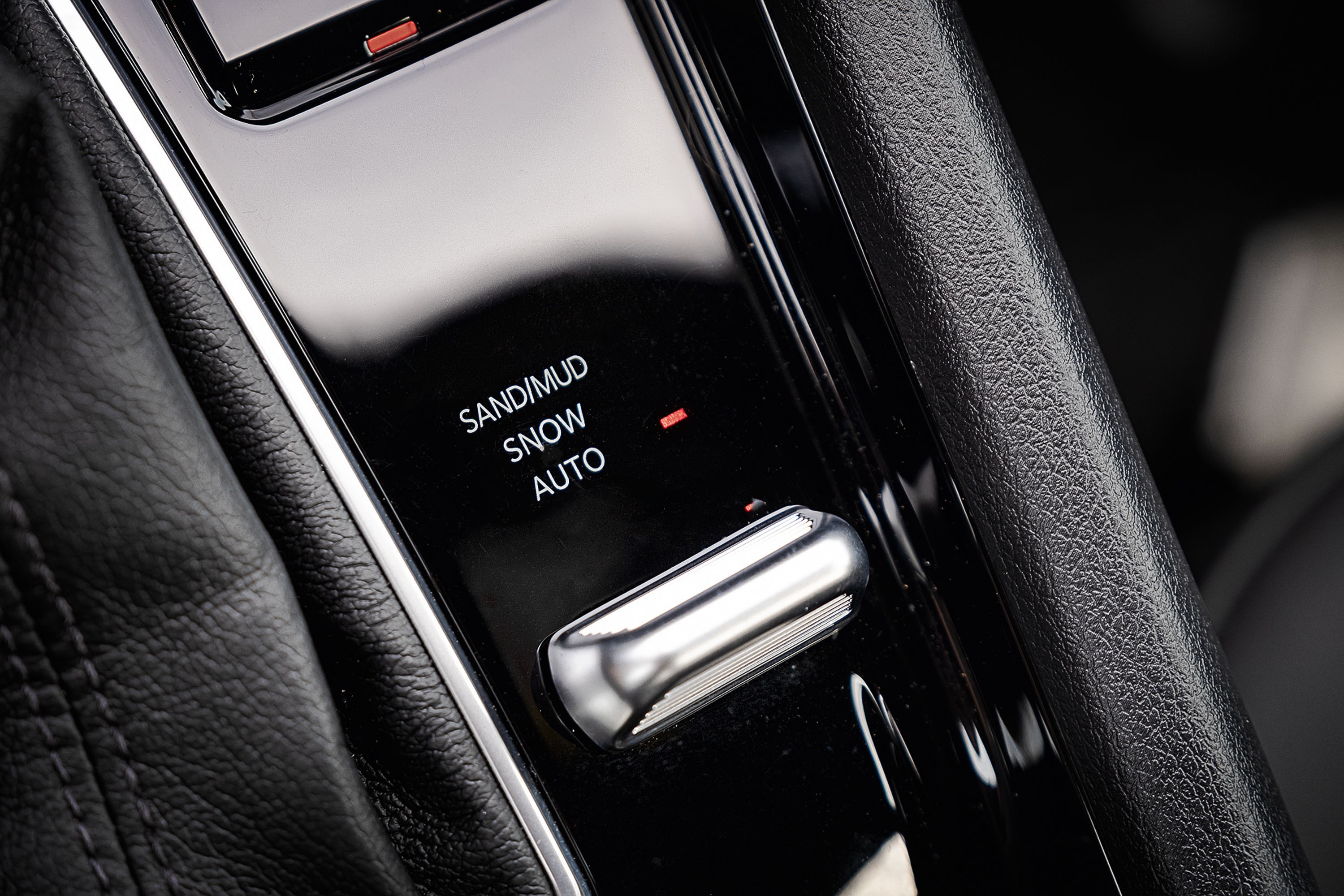Painel de controle de carro com seletor de modo de condu&ccedil;&atilde;o. As op&ccedil;&otilde;es vis&iacute;veis s&atilde;o SAND/MUD, SNOW e AUTO, com um indicador vermelho ao lado de SNOW. Um bot&atilde;o cromado alongado est&aacute; abaixo das op&ccedil;&otilde;es, e o painel &eacute; preto brilhante, ladeado por estofamento de couro preto texturizado