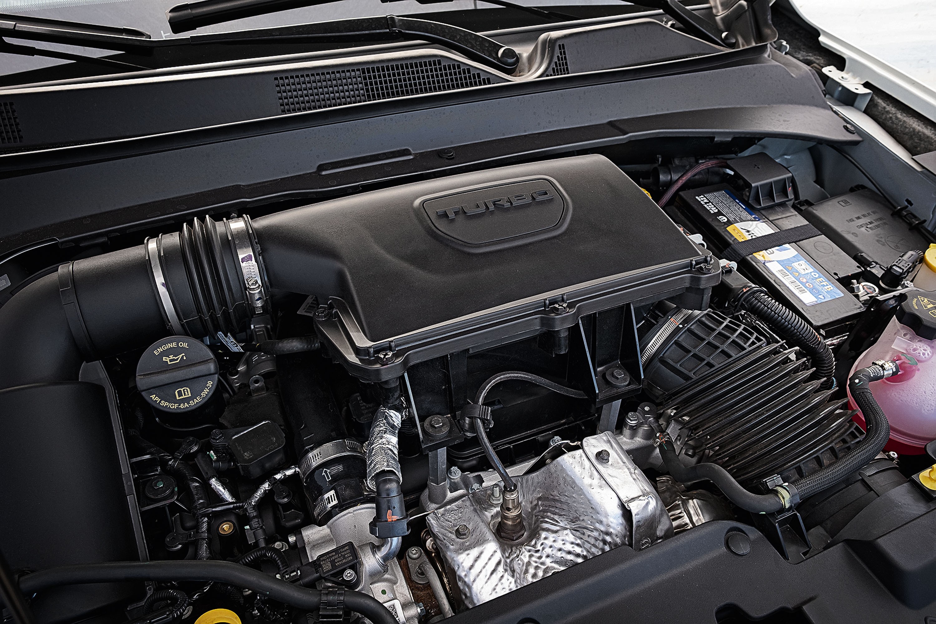 Vista superior do motor de um carro, com a tampa preta do motor exibindo a palavra TURBO em relevo. &Agrave; esquerda, o bocal de &oacute;leo com o s&iacute;mbolo de uma almotolia. &Agrave; direita, a bateria do ve&iacute;culo e o reservat&oacute;rio de fluido rosa. Diversos tubos, fios e componentes met&aacute;licos prateados est&atilde;o vis&iacute;veis, com o para-brisa e a grade de ventila&ccedil;&atilde;o do carro ao fundo