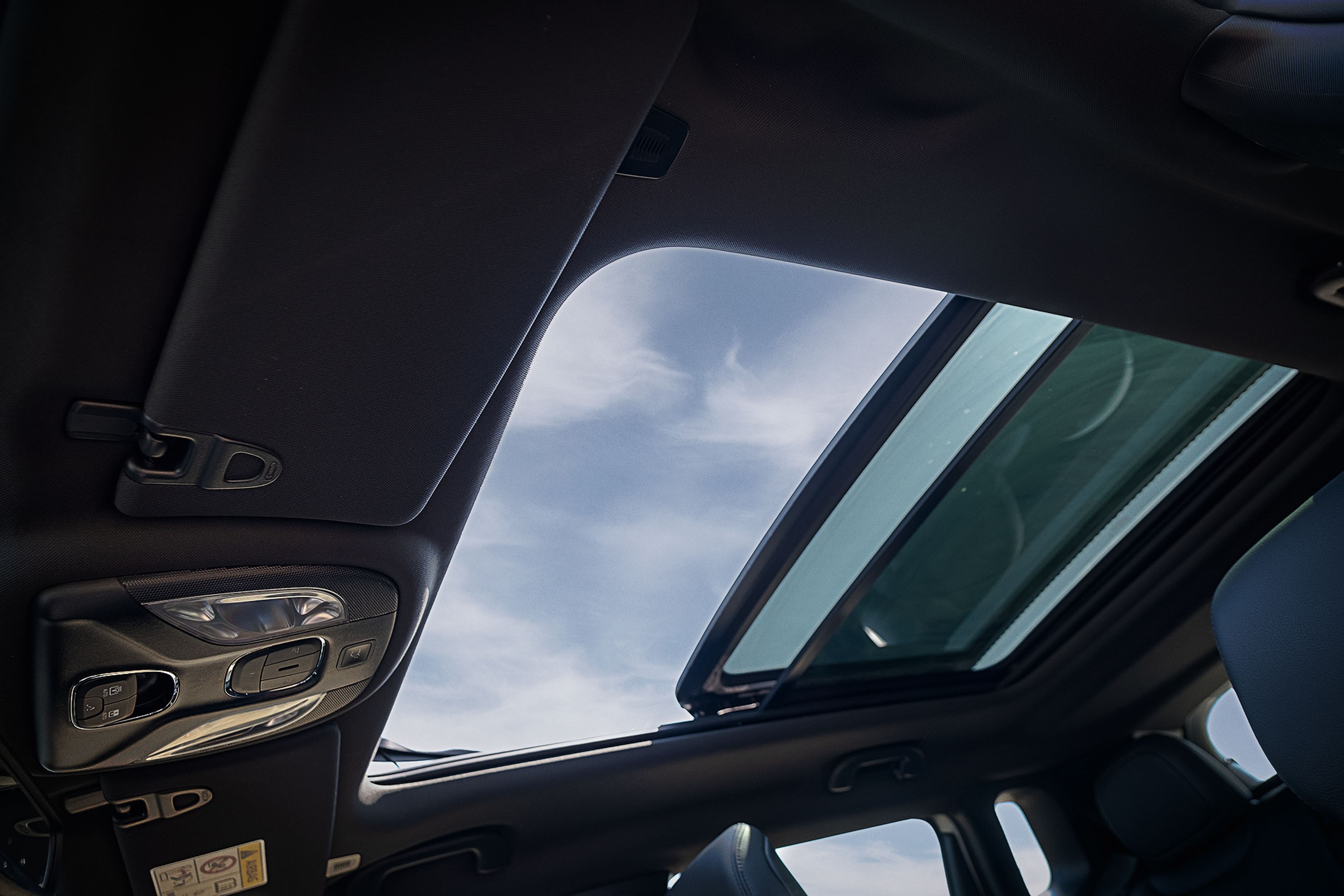 Vista interna do teto de um carro com teto solar panor&acirc;mico aberto, revelando um c&eacute;u azul com nuvens esparsas. O interior &eacute; escuro, com detalhes do console de luzes e para-sol vis&iacute;veis
