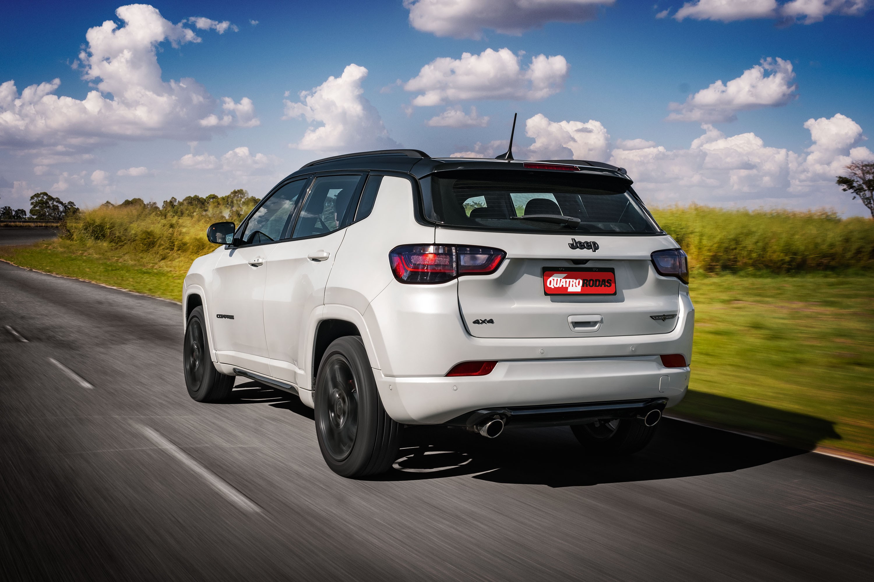 SUV Jeep Compass branco com teto preto, visto por tr&aacute;s e lateral, em movimento numa estrada asfaltada. O c&eacute;u azul tem nuvens brancas e h&aacute; vegeta&ccedil;&atilde;o verde e amarelada nas margens