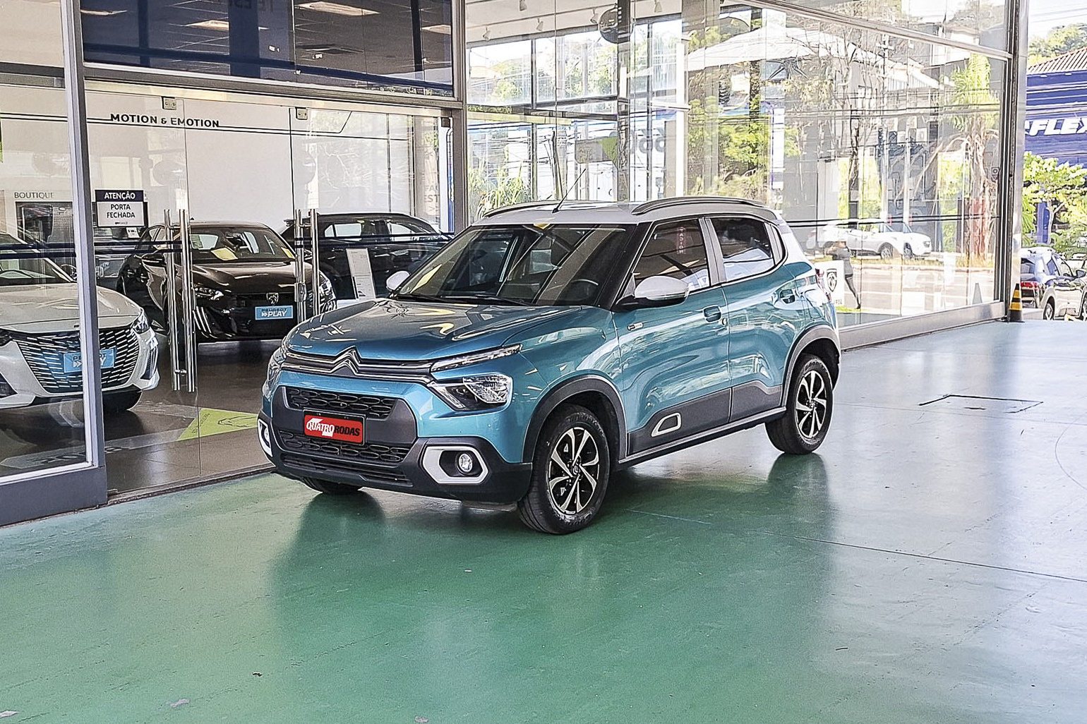 Um carro SUV Citroën C3 azul-turquesa com teto branco está estacionado em uma concessionária, com outros carros visíveis através da vitrine de vidro. O piso é verde-claro e reflete a luz.