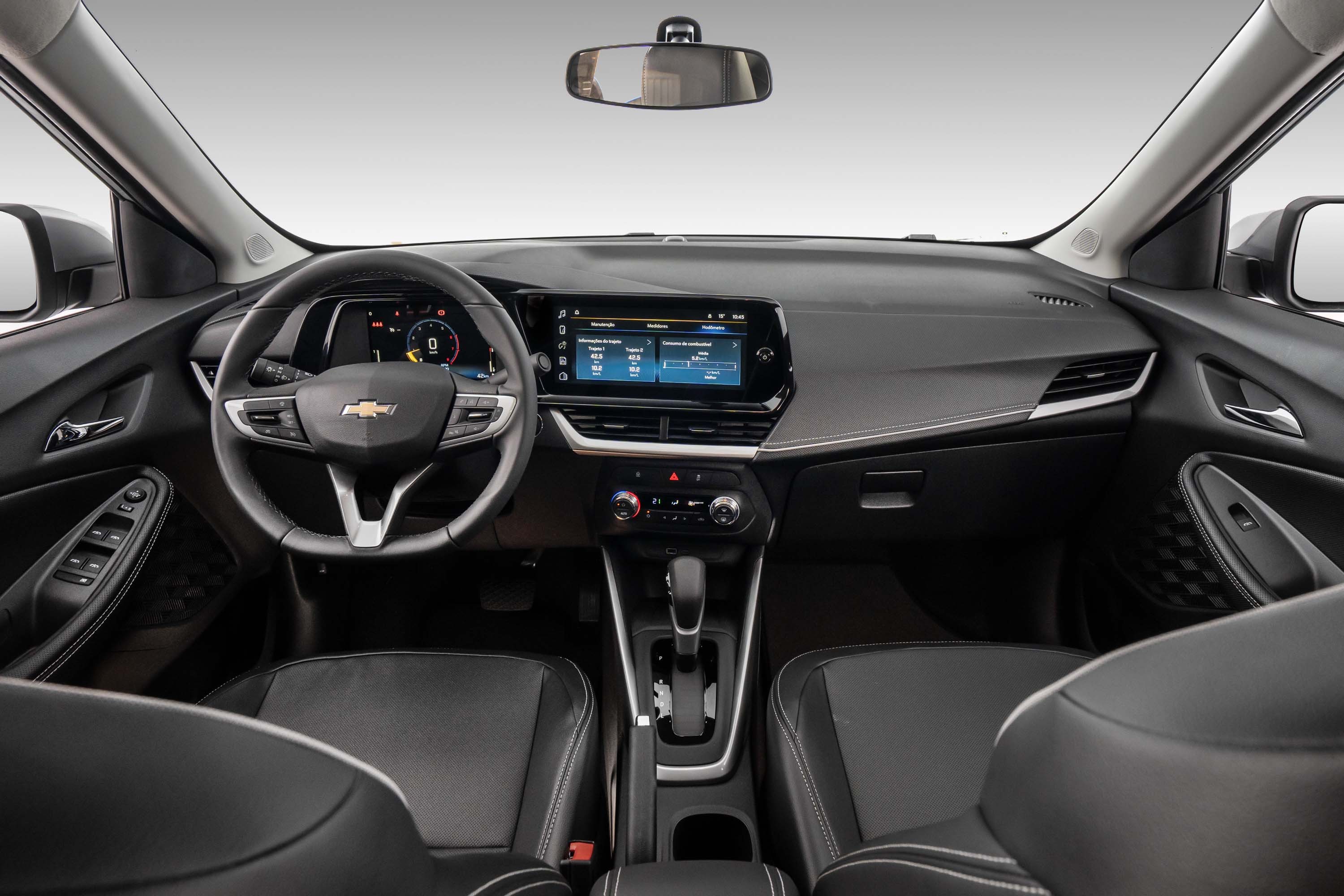 Interior de um carro moderno, com painel em tons de cinza escuro e detalhes prateados. O volante multifuncional com o logo da Chevrolet est&aacute; &agrave; esquerda, e uma tela multim&iacute;dia digital exibe informa&ccedil;&otilde;es no centro. Os bancos dianteiros s&atilde;o pretos com costuras brancas, e o c&acirc;mbio autom&aacute;tico est&aacute; vis&iacute;vel no console central. O espelho retrovisor interno reflete o teto do ve&iacute;culo