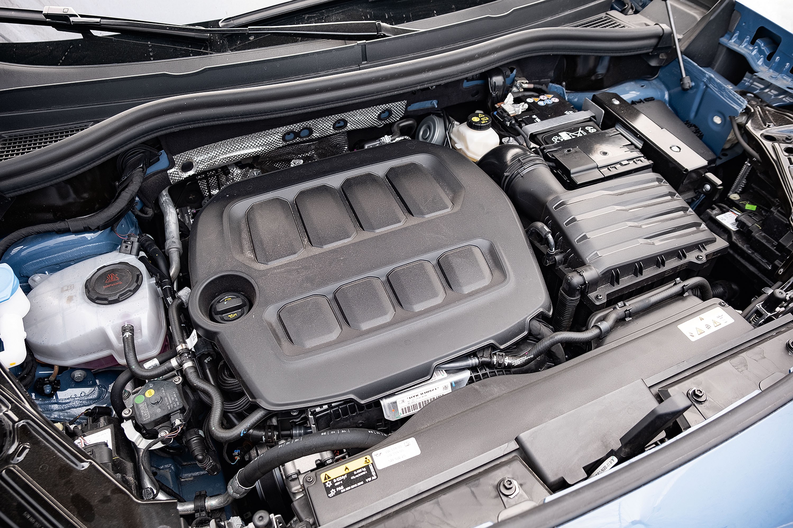 Vista superior do motor de um carro, com a tampa do motor preta em destaque no centro, cercada por mangueiras, reservat&oacute;rios e componentes met&aacute;licos e pl&aacute;sticos, em um compartimento azul