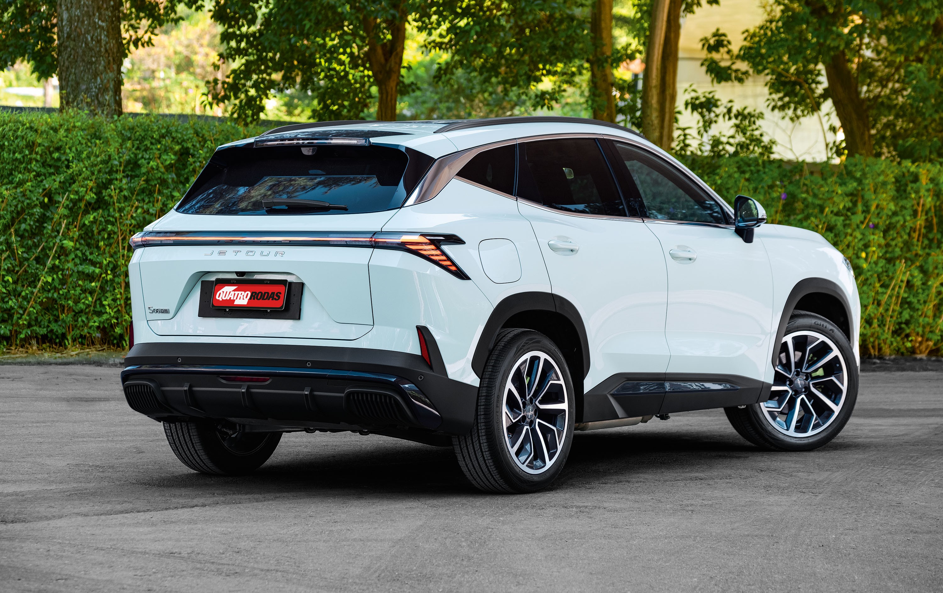 SUV Jetour branco, visto de traseira e lateral, estacionado em asfalto cinza com vegeta&ccedil;&atilde;o verde ao fundo. A placa traseira vermelha exibe QUATRO RODAS