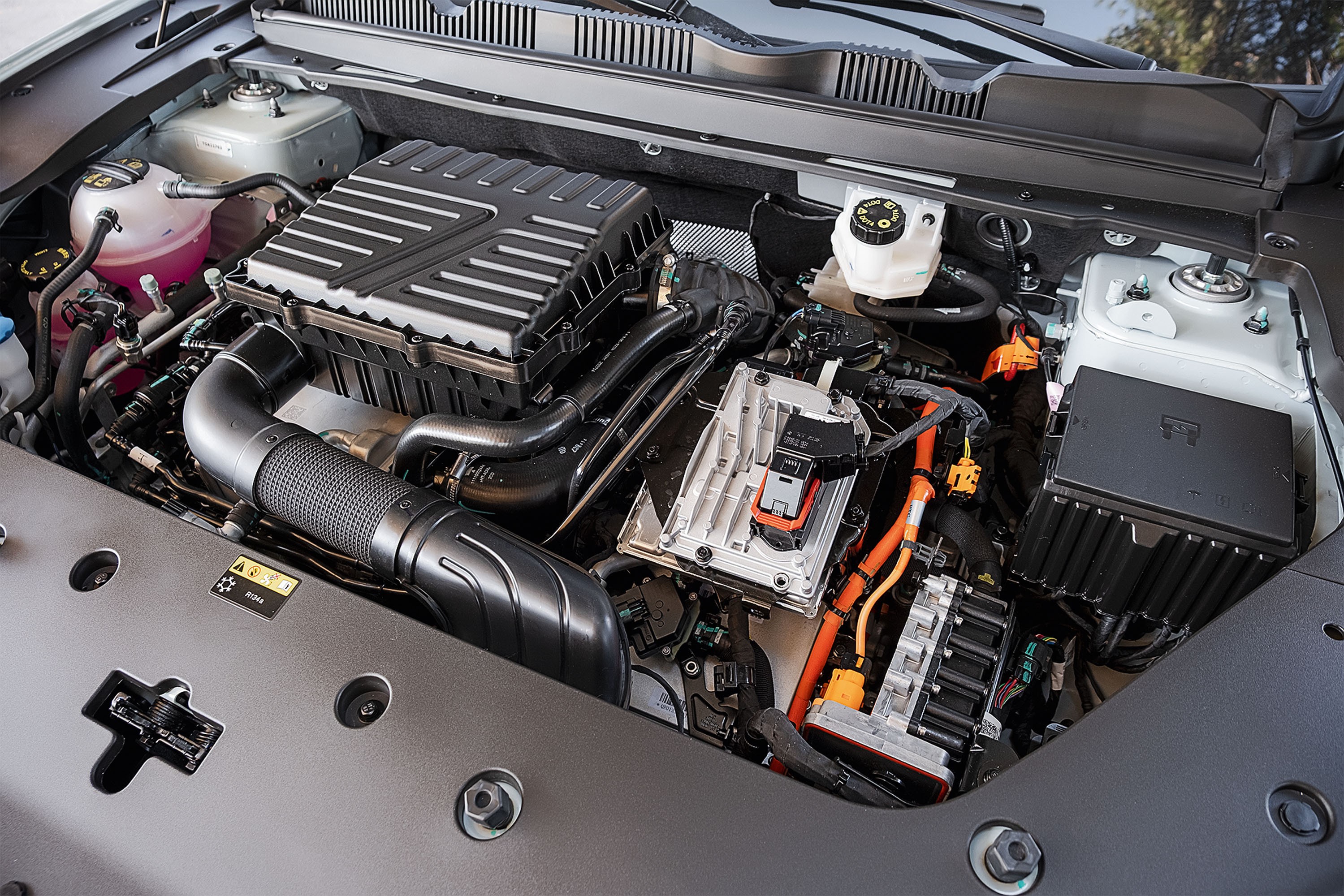 Motor de carro h&iacute;brido com componentes el&eacute;tricos e mec&acirc;nicos. Destacam-se cabos laranja, reservat&oacute;rios de fluidos, caixa de fus&iacute;veis preta e um grande filtro de ar.