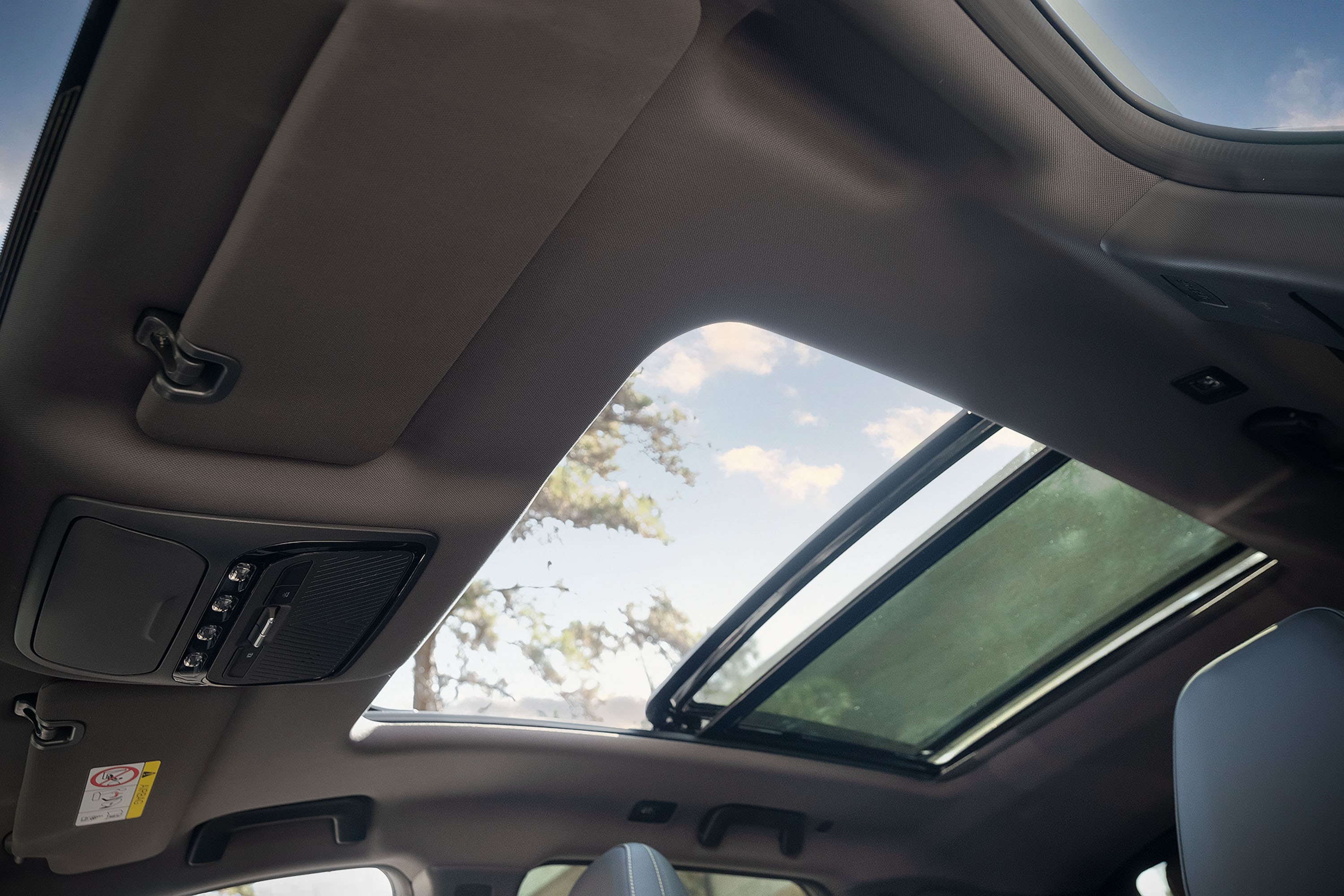 Interior de um carro com teto solar panor&acirc;mico aberto, mostrando o c&eacute;u azul com nuvens e &aacute;rvores ao fundo. O forro do teto &eacute; cinza, com para-sol e console de luzes e bot&otilde;es