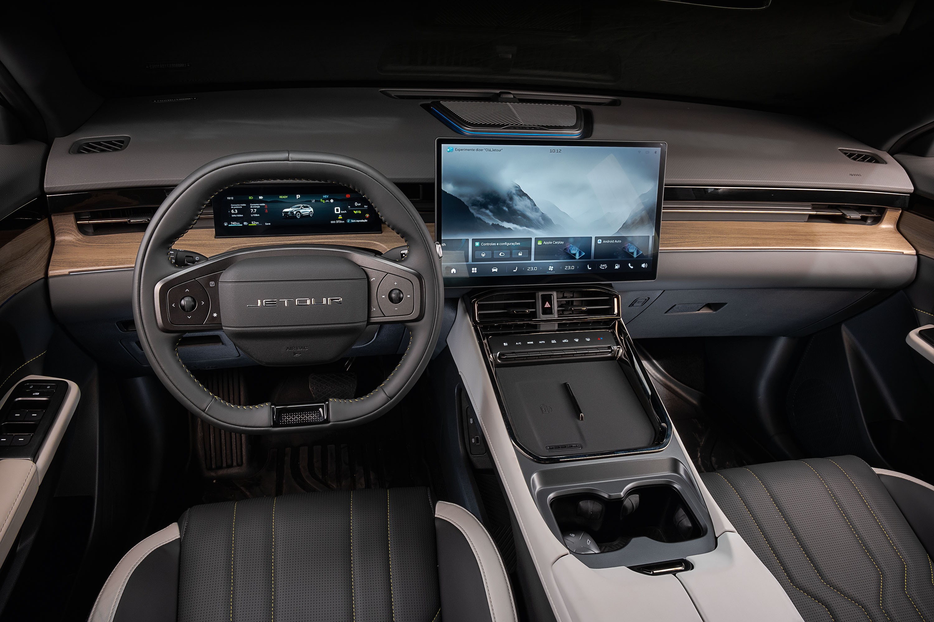 Interior moderno de um carro, com volante preto e o nome JETOUR no centro. O painel possui uma tela digital exibindo um carro e outra tela maior no centro com uma paisagem montanhosa e op&ccedil;&otilde;es de menu. Detalhes em madeira clara e acabamentos cinza escuro e prata complementam o design, com bancos de couro preto e costura amarela vis&iacute;veis no canto inferior direito