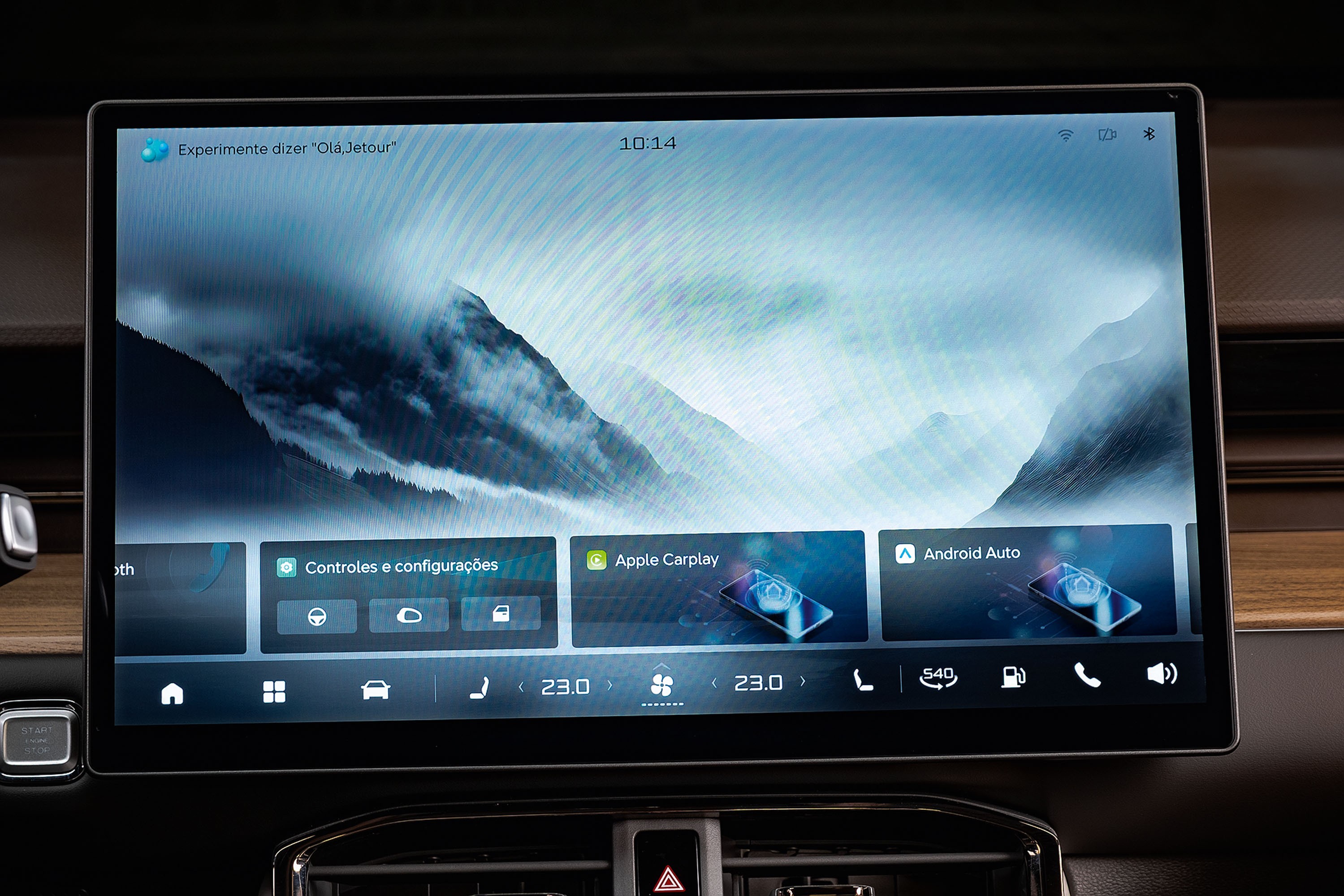 Tela multim&iacute;dia de carro exibindo um papel de parede de montanhas nebulosas. Na parte inferior, &iacute;cones de navega&ccedil;&atilde;o, controles do ve&iacute;culo, Apple CarPlay e Android Auto. No topo, a hora 10:14 e a sugest&atilde;o Experimente dizer Ol&aacute; Jetour.