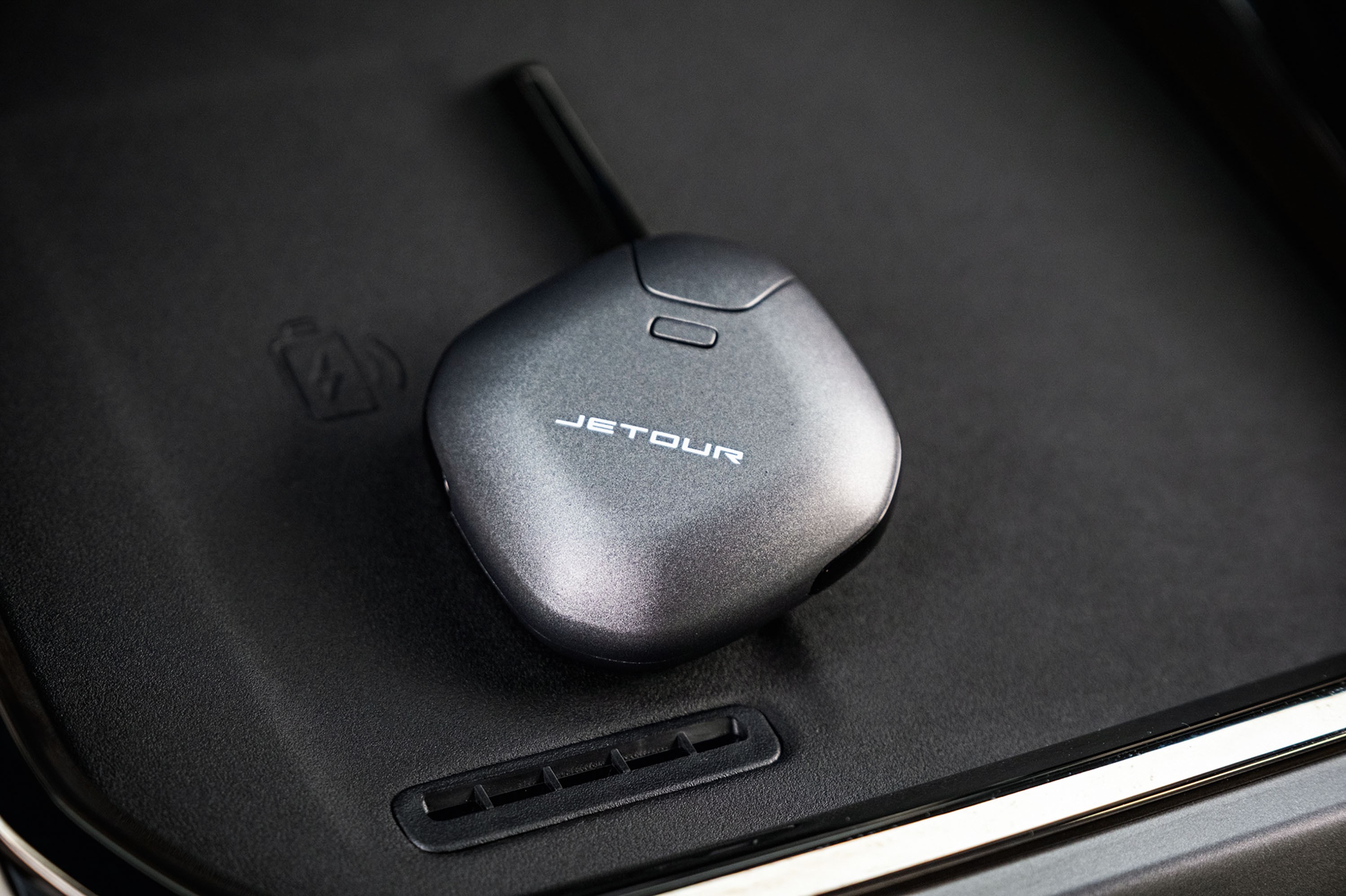 Chave de carro Jetour cinza-chumbo com antena preta, repousando sobre um console preto texturizado de ve&iacute;culo, com um &iacute;cone de carregamento sem fio em relevo